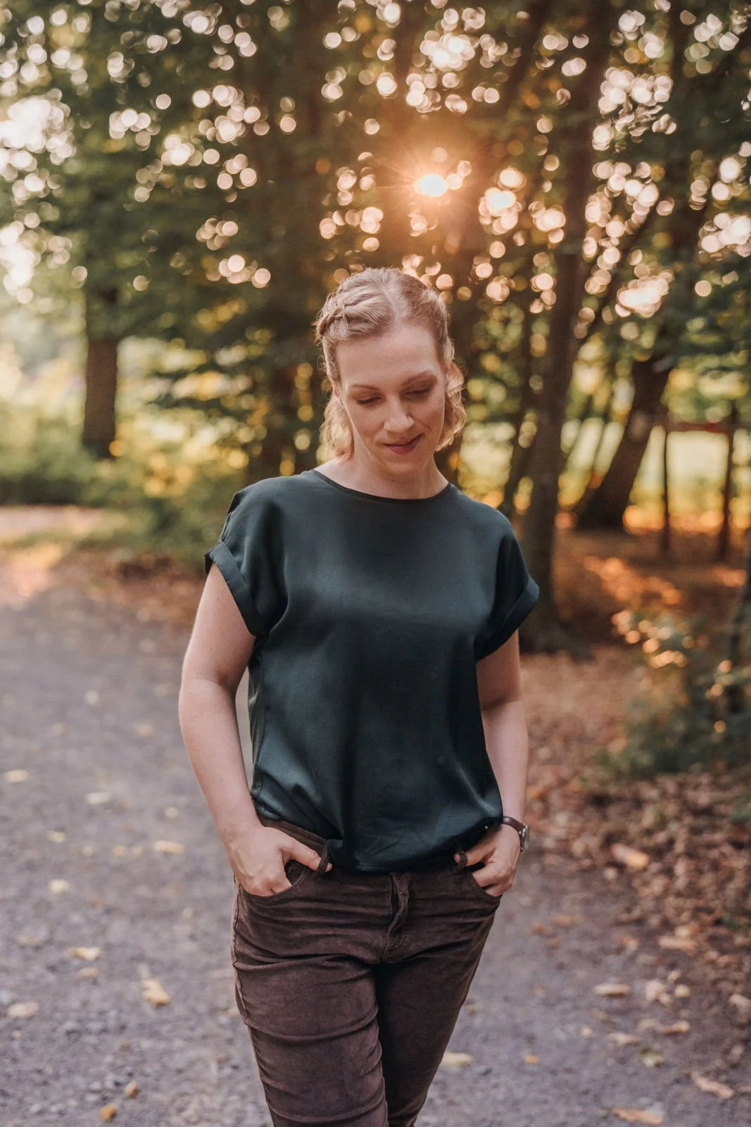 Junge Frau geht im Park im Wald bei Sonnenuntergang, trägt schwarze Bluse und braune Hosen.
