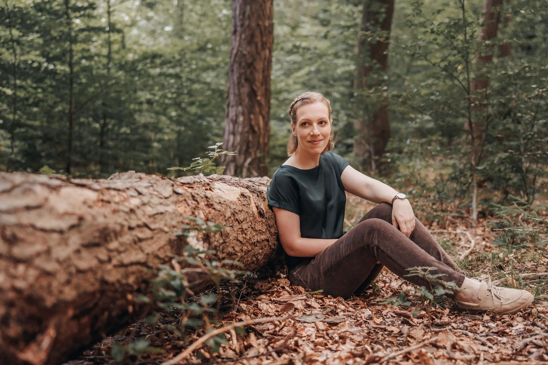 Junge Frau sitzt im Wald neben einem gefallenen Baum, umgeben von Blättern und Bäumen.