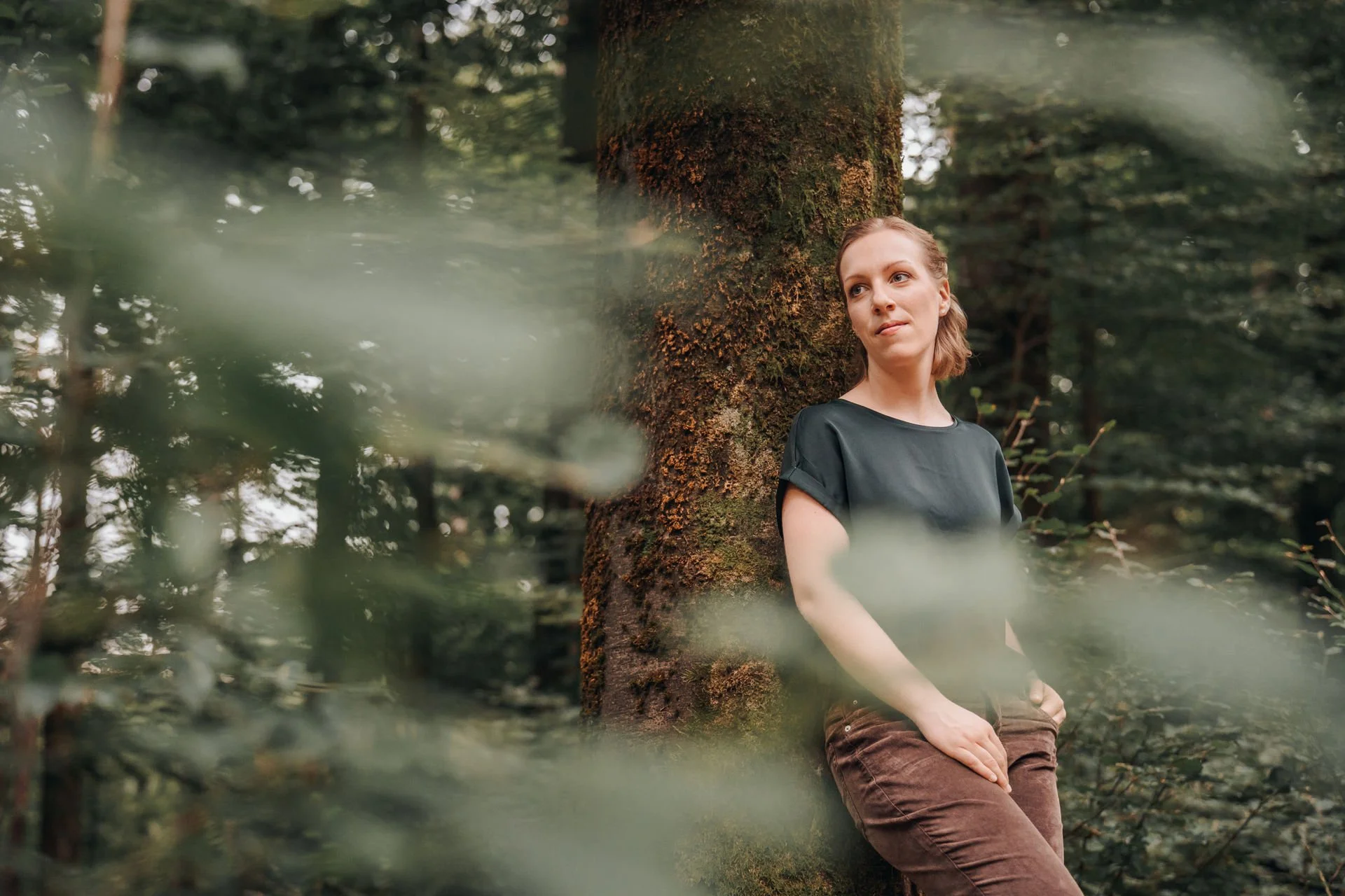 Junge Frau lehnt an einem Baum im Wald, während sie nach rechts schaut.