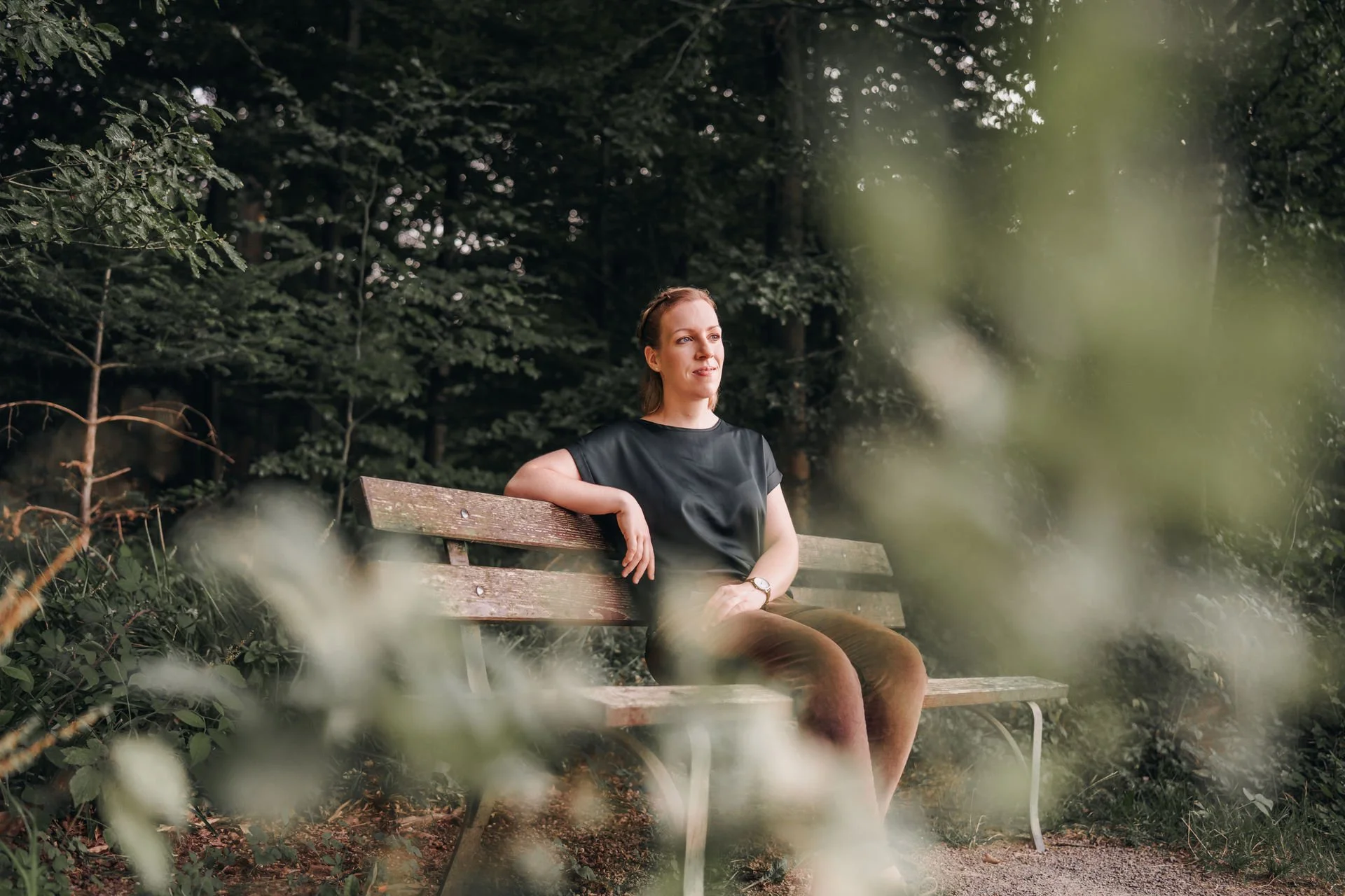 Eine junge Frau sitzt entspannt auf einer Parkbank im Wald, umgeben von Büschen und Bäumen.