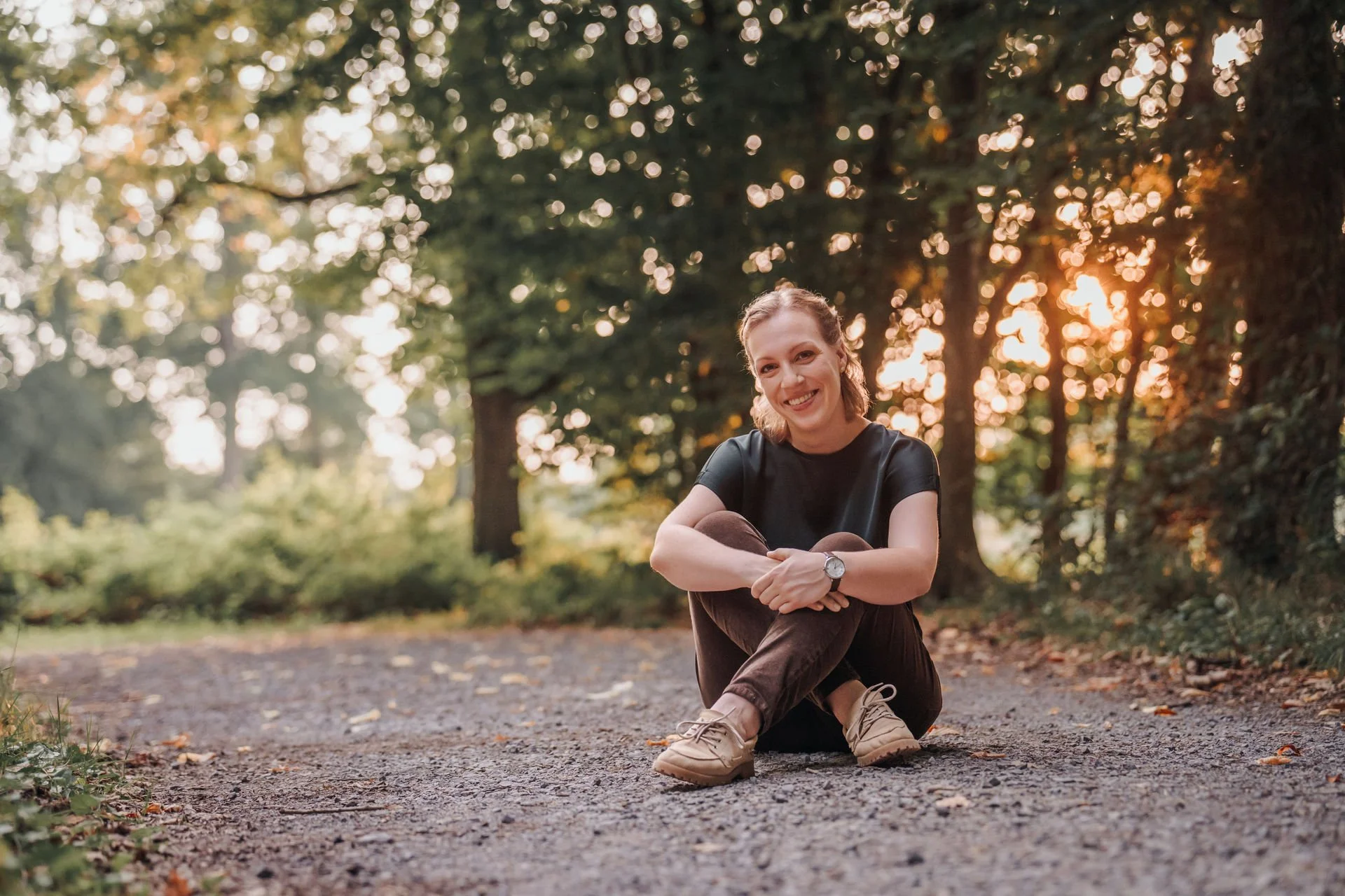 Eine lachende junge Frau sitzt auf einem Weg bei Sonnenuntergang