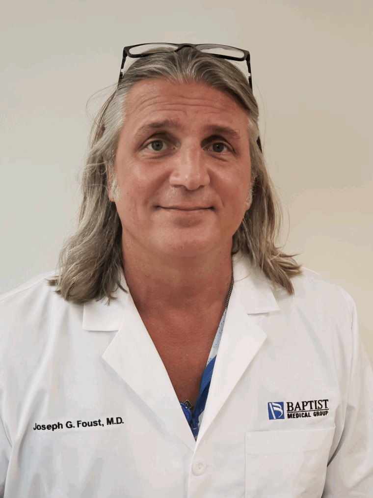 A middle-aged man with long gray hair, wearing glasses on his head, smiling, in a white medical coat with 'Joseph G. Foust, M.D.' embroidered on it, and a Baptist Medical Group logo.