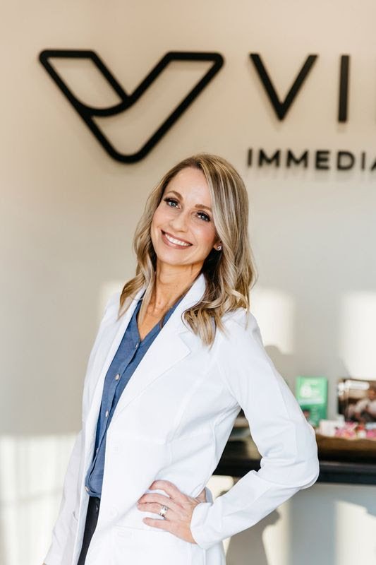Female doctor in white coat with stethoscope around neck smiling outdoors near a brick wall.