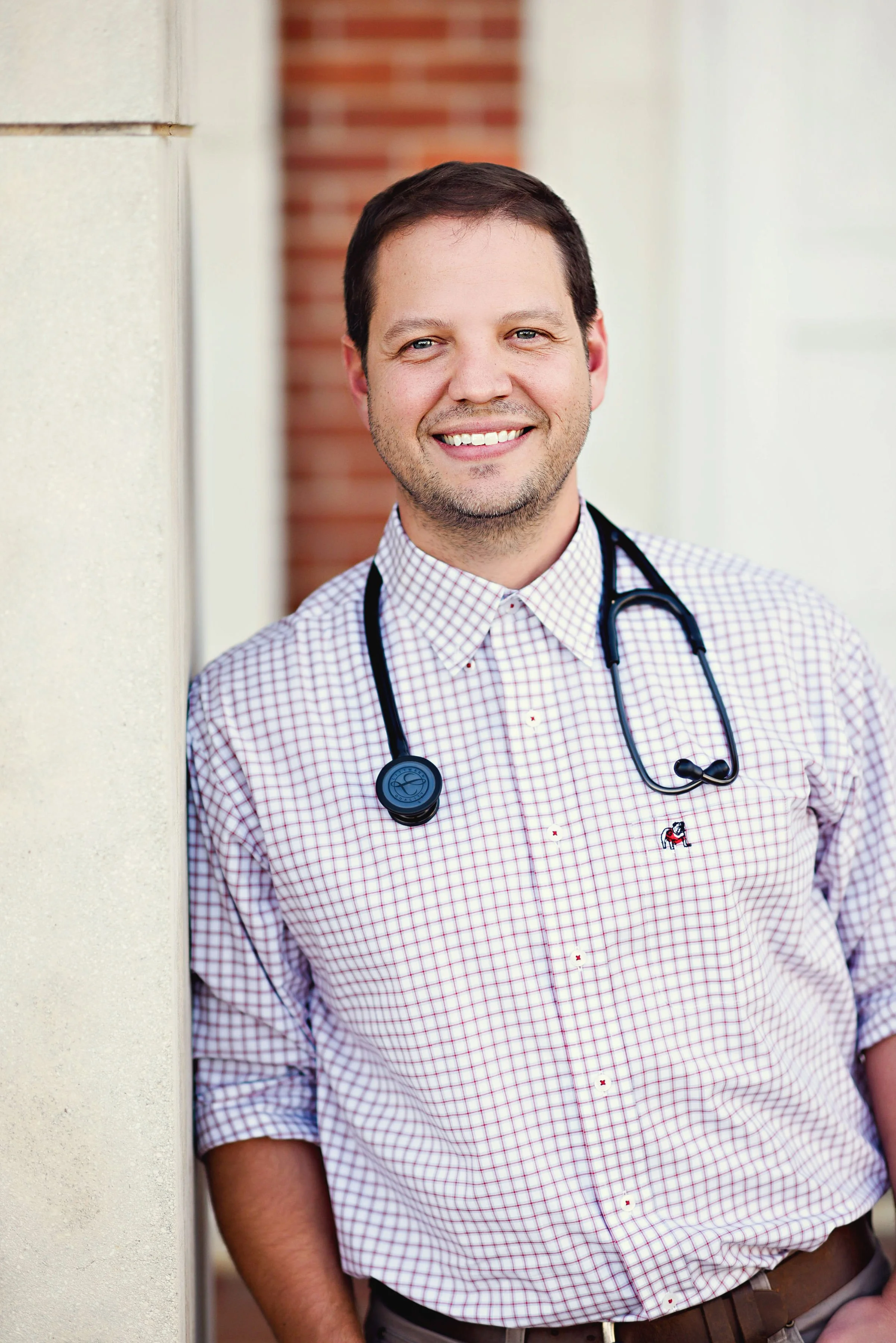 A smiling male doctor leaning against a wall outdoors, wearing a checked shirt with a stethoscope around his neck.