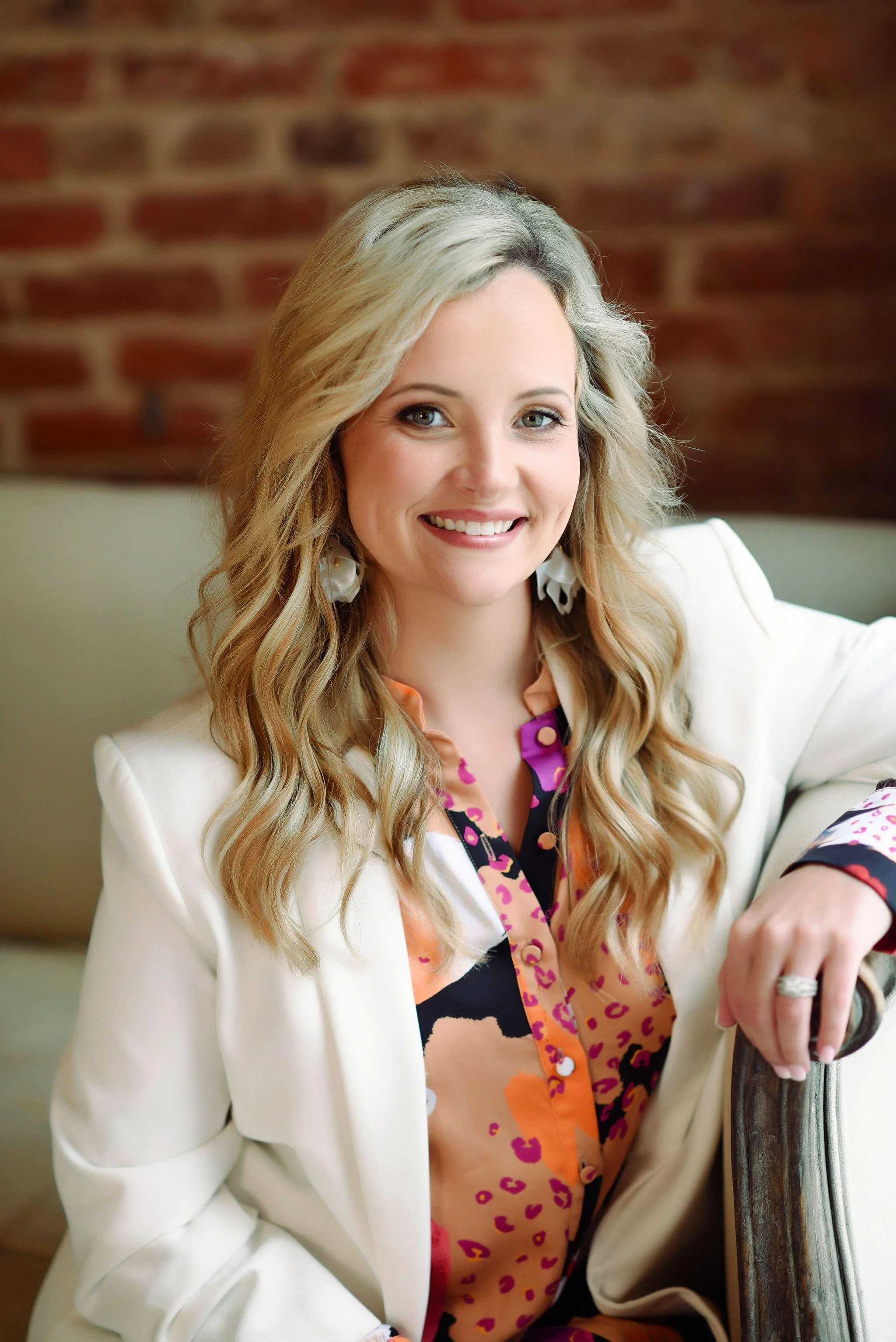 A smiling woman with long, wavy blonde hair, wearing a white blazer over a colorful, patterned blouse, sitting on a light-colored sofa with a brick wall in the background.