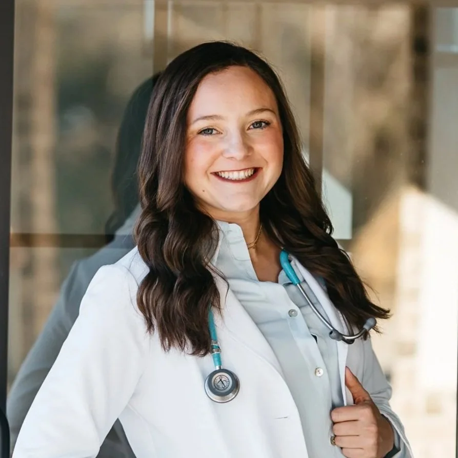 A smiling female doctor in a white coat with a stethoscope around her neck, standing indoors.