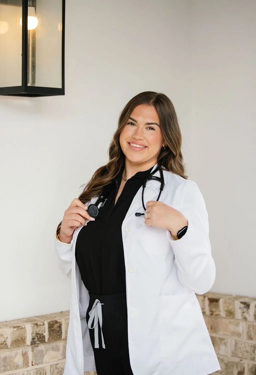 Female doctor in white coat with stethoscope around neck smiling outdoors near a brick wall.