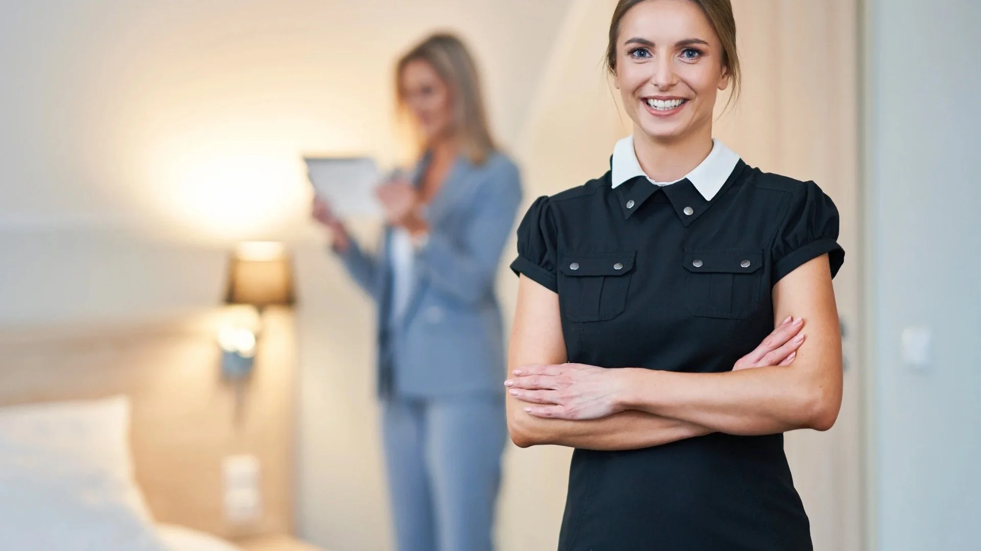 Due donne in un ambiente domestico, una in primo piano con abiti scuri e un sorriso, l'altra sullo sfondo con abiti chiari che utilizza un tablet.