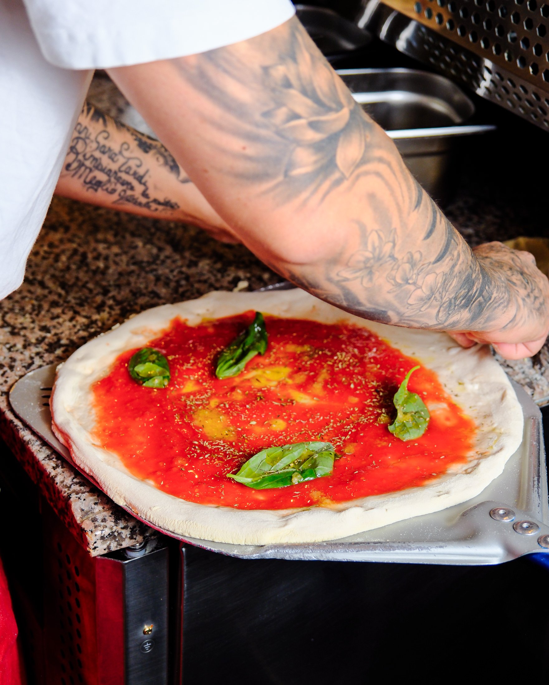A person with tattoos preparing to bake a pizza with tomato sauce, basil leaves, and spices.