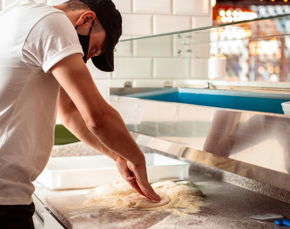 A man wearing a black cap and a white t-shirt making pizza dough on a floured countertop in a pizzeria kitchen.