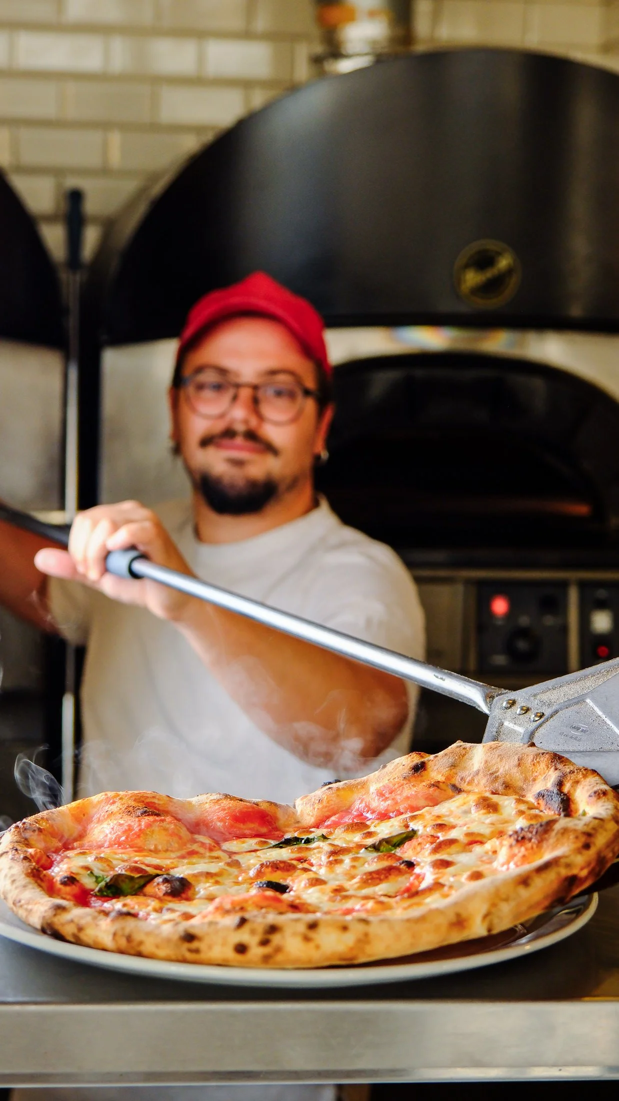 A man with glasses and a red cap is holding a pizza peel with a freshly baked pizza, in front of a pizza oven.