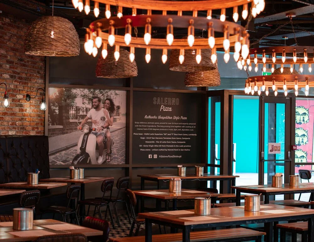 Salerno Pizza restaurant interior showing exposed brick walls and wooden tables set with placemats and metallic containers. Modern chandelier and wicker pendant lights hang from the ceiling.