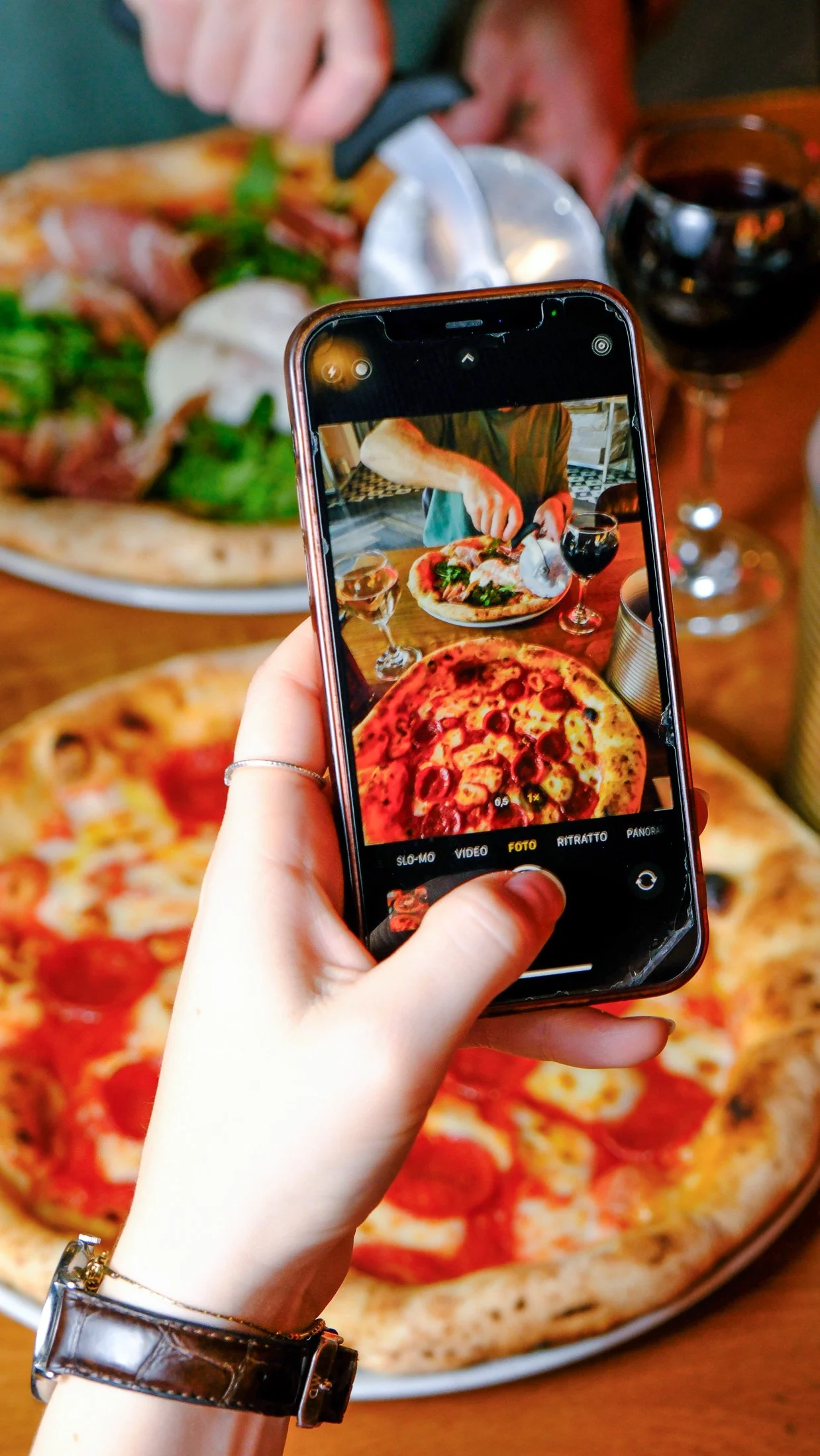 A person taking a photo of a pizza with a smartphone; the pizza has pepperoni slices and cheese, and there are glasses of red wine on the table.