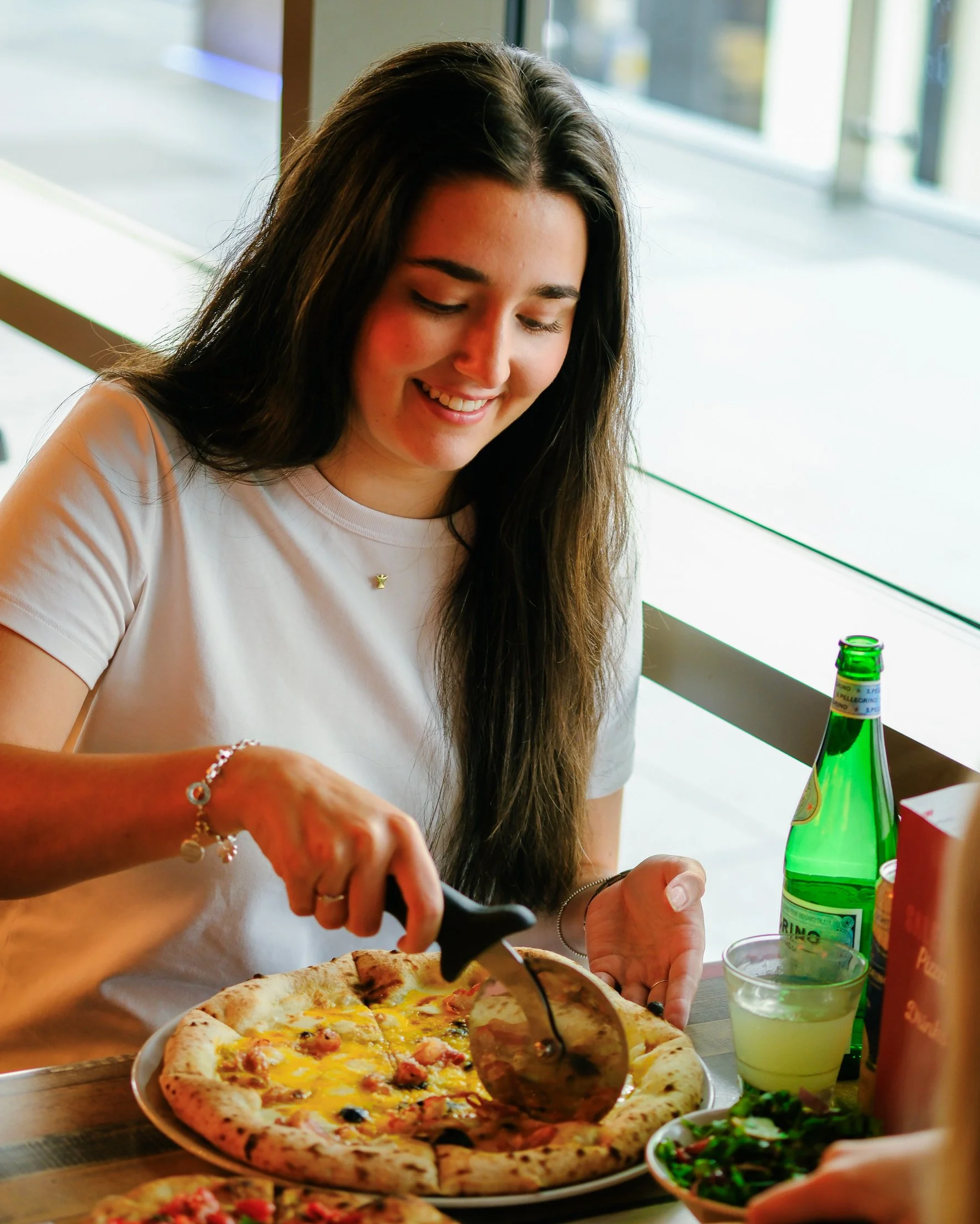 A young woman with long brown hair smiling while slicing a pizza at a dining table in a restaurant, with a green bottle, a glass of soda, and a salad nearby.