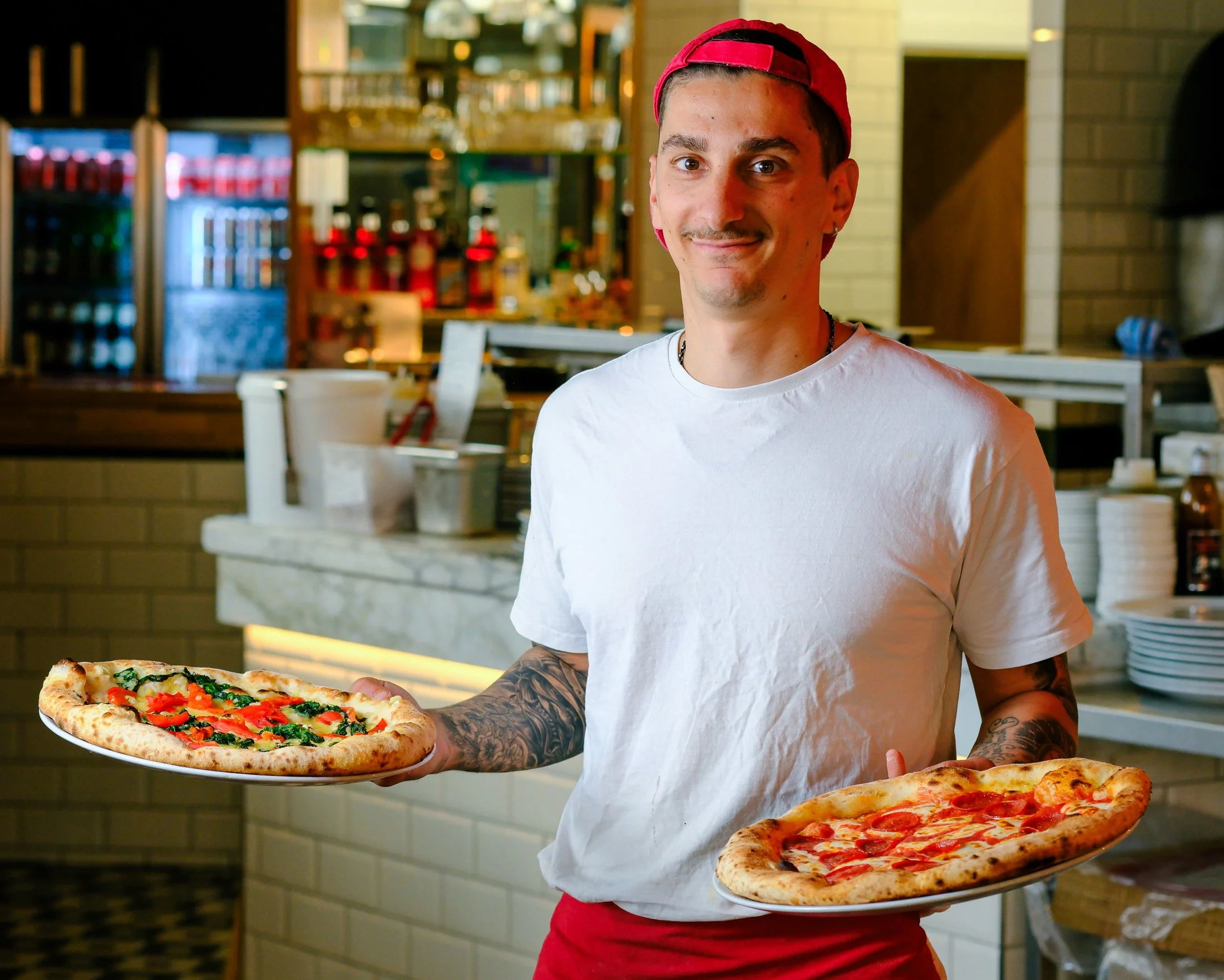 A smiling man with tattoos holds two pizza plates inside a pizzeria. One pizza has vegetables and the other has pepperoni.
