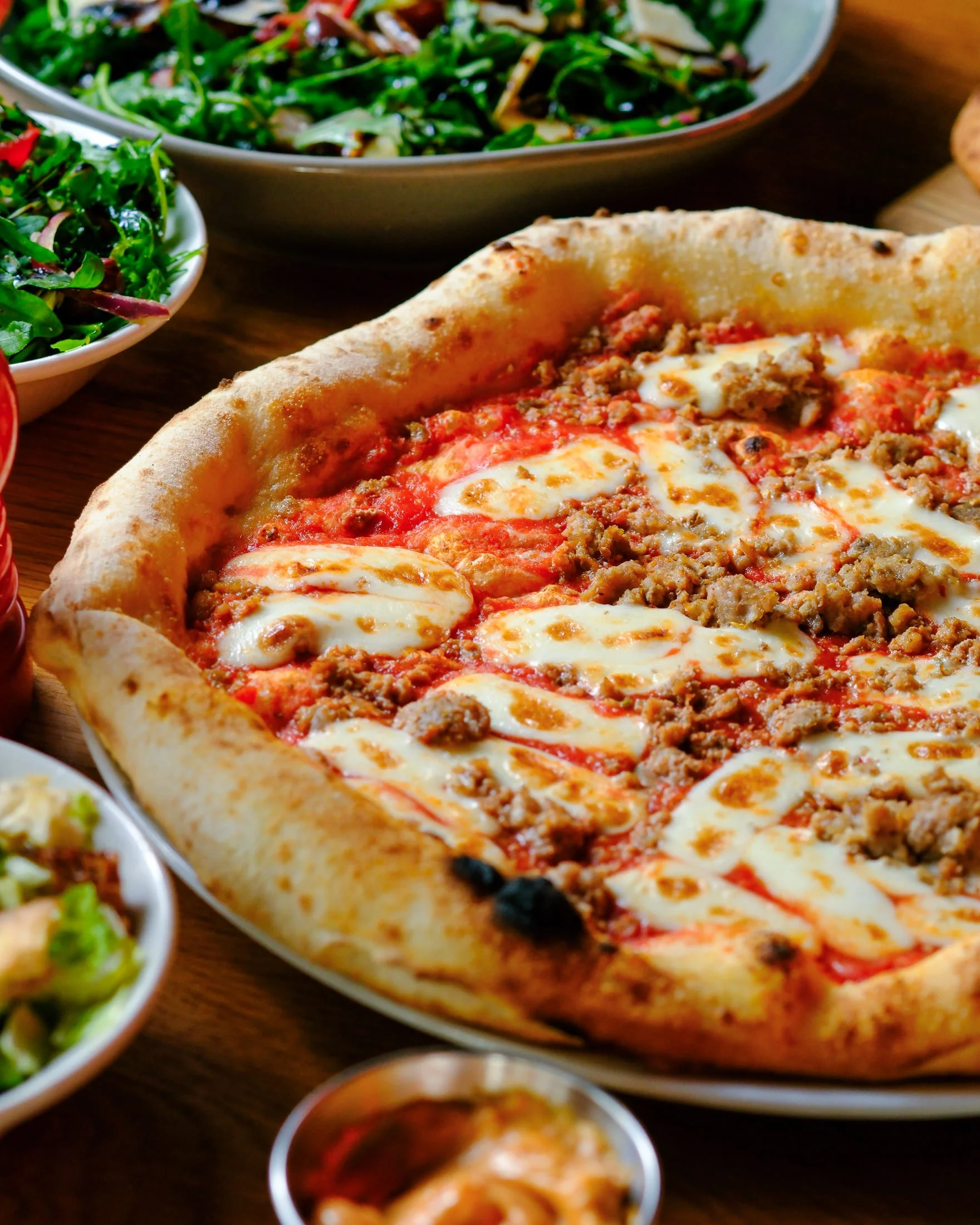 A young woman with long brown hair smiling while slicing a pizza at a dining table in a restaurant, with a green bottle, a glass of soda, and a salad nearby.