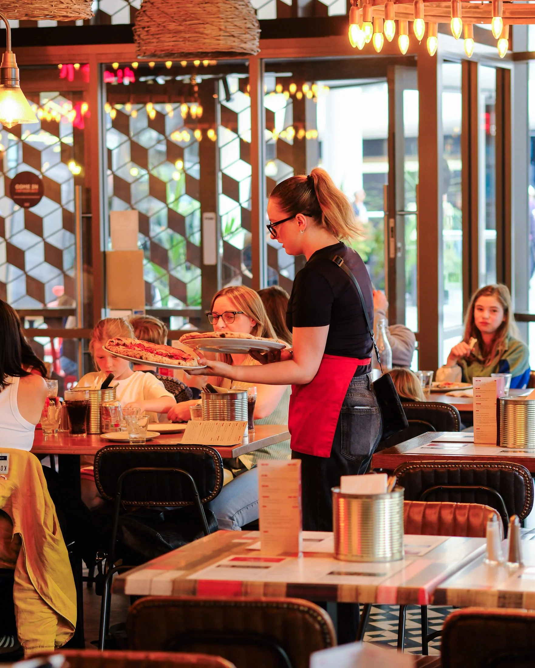 A waitress carrying two plates of pizza in a busy restaurant with children and adults dining at tables, large windows, and decorative lighting.