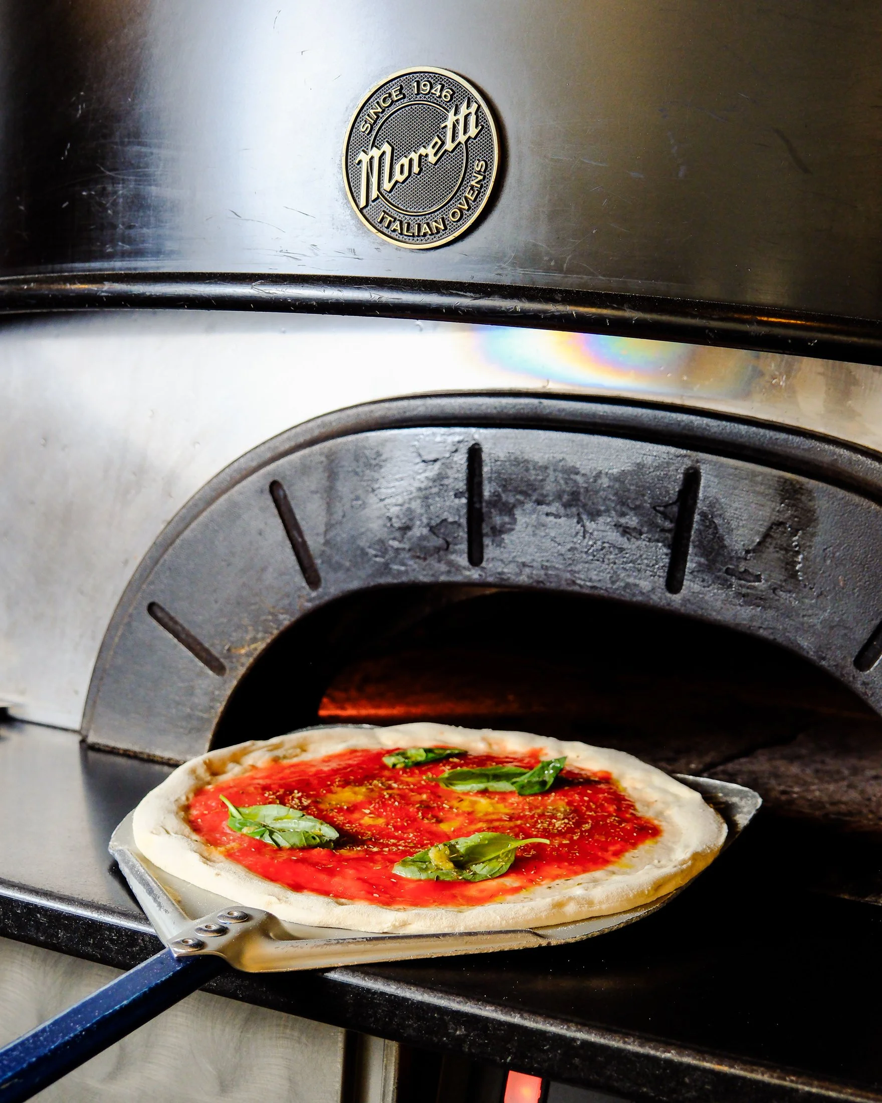 A pizza being cooked in a wood-fired oven with a metallic exterior and a logo that reads "Mariotti Italian Ovens".