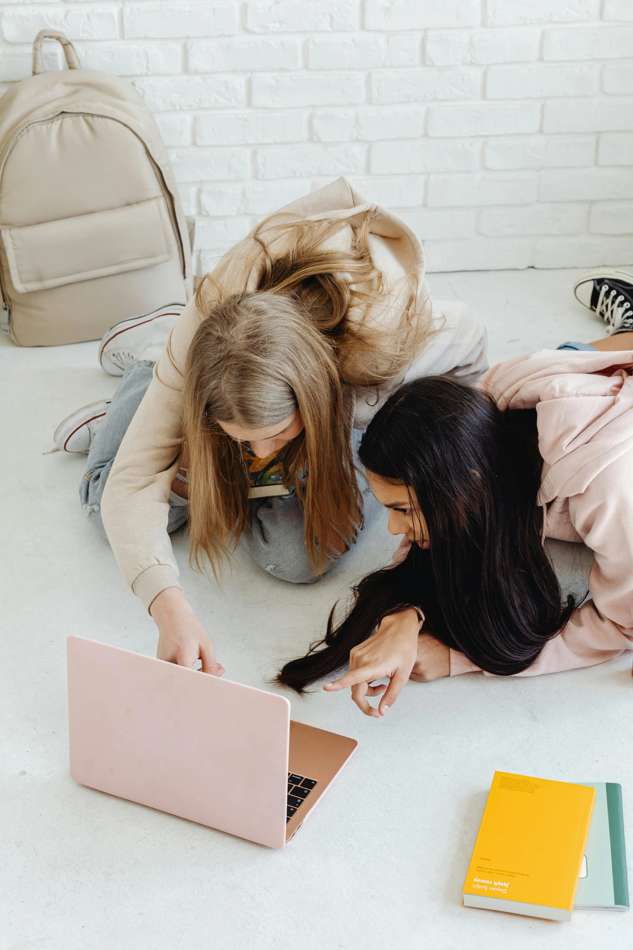 Two girls lying on the floor looking at a pink laptop, with a yellow notebook nearby, in front of a white brick wall and a beige backpack.