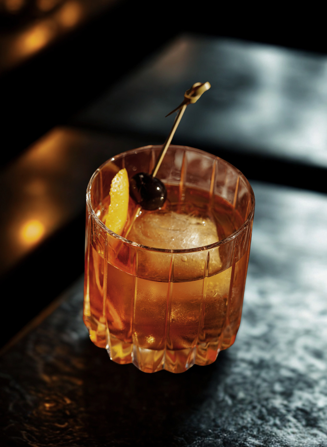 A cocktail in a short, textured glass garnished with a lemon wedge and a dark cherry, with a large ice cube inside.