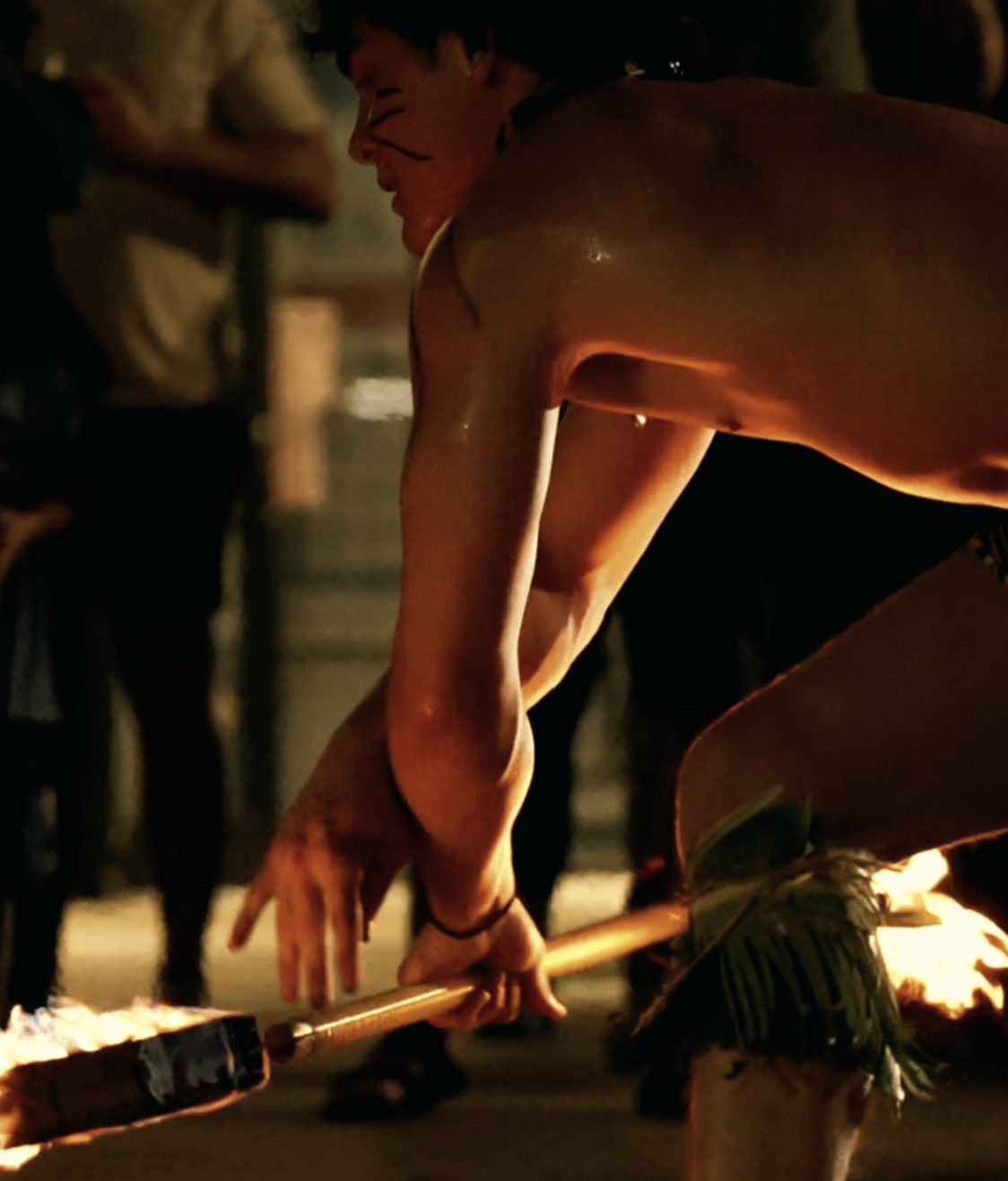 A person crouching and smiling, holding a flaming torch, with other people blurred in the background.