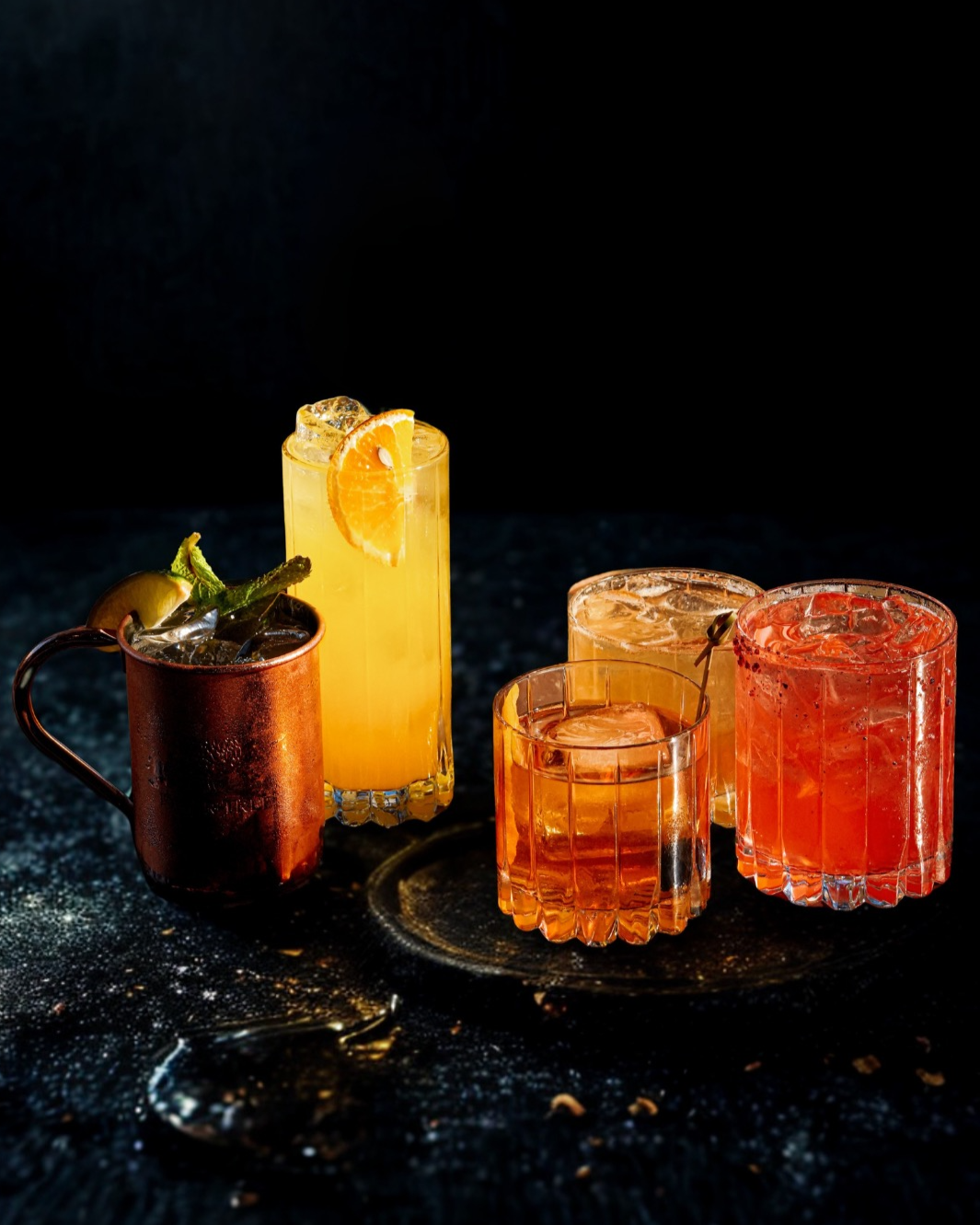 Five colorful cocktails with ice on a dark surface, featuring a mug with a lime wedge, a tall glass with orange slice, two short glasses with reddish drinks, and a small glass with clear drink.