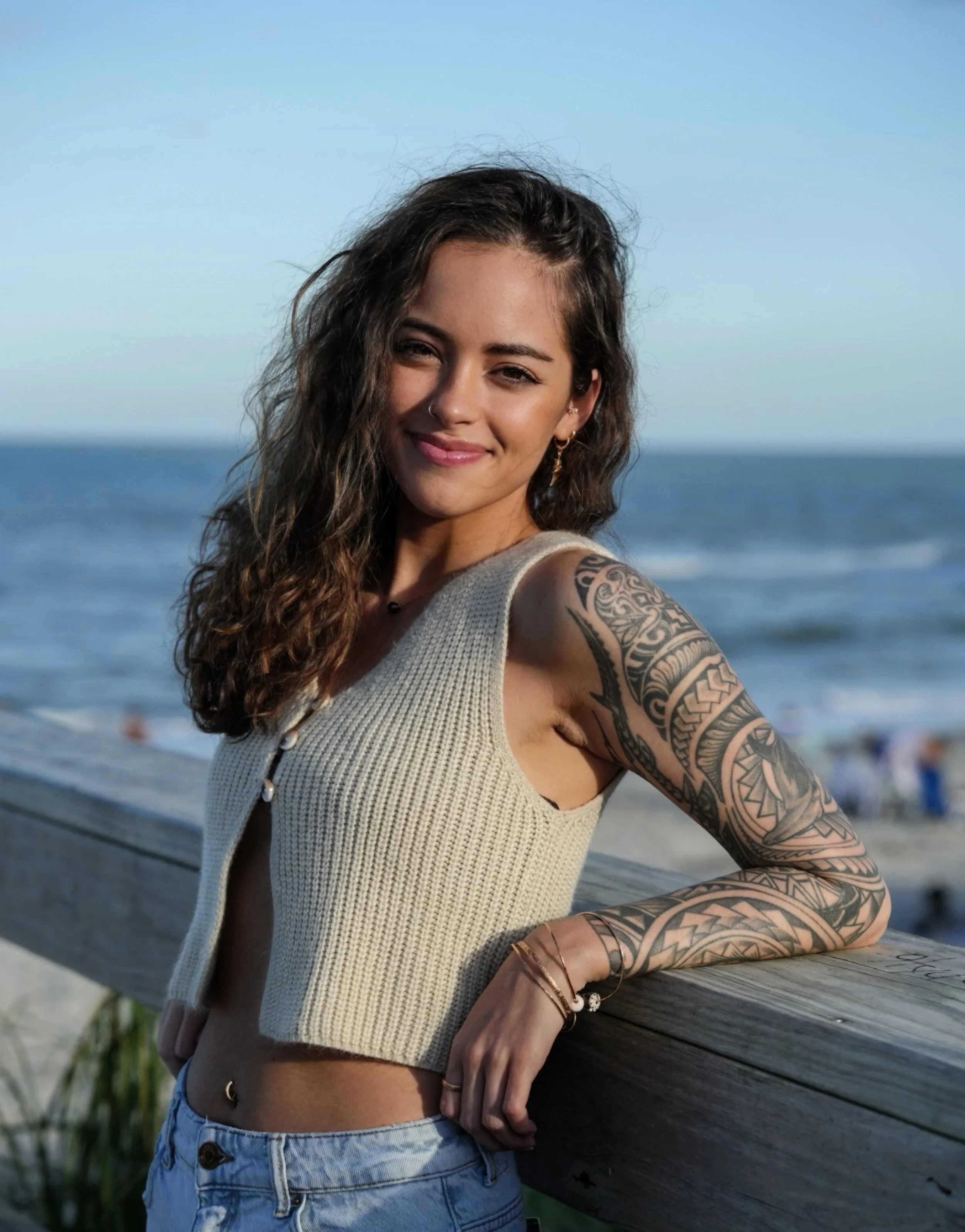 Young woman with tattoos on her arm leaning on a wooden railing at the beach, smiling, with ocean and sky in the background.