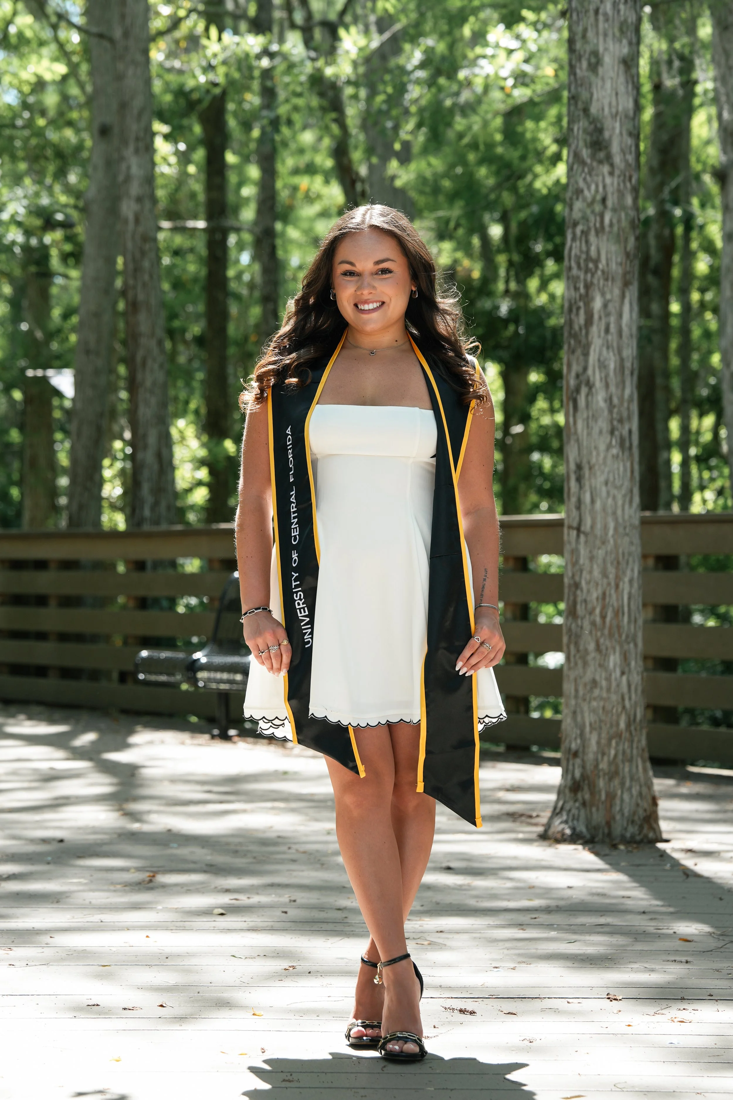 A young woman in a white dress and black high heels stands outdoors on a wooden deck, wearing a black graduation stole with yellow trim that reads "University of Central Florida," surrounded by tall trees and a wooden fence.
