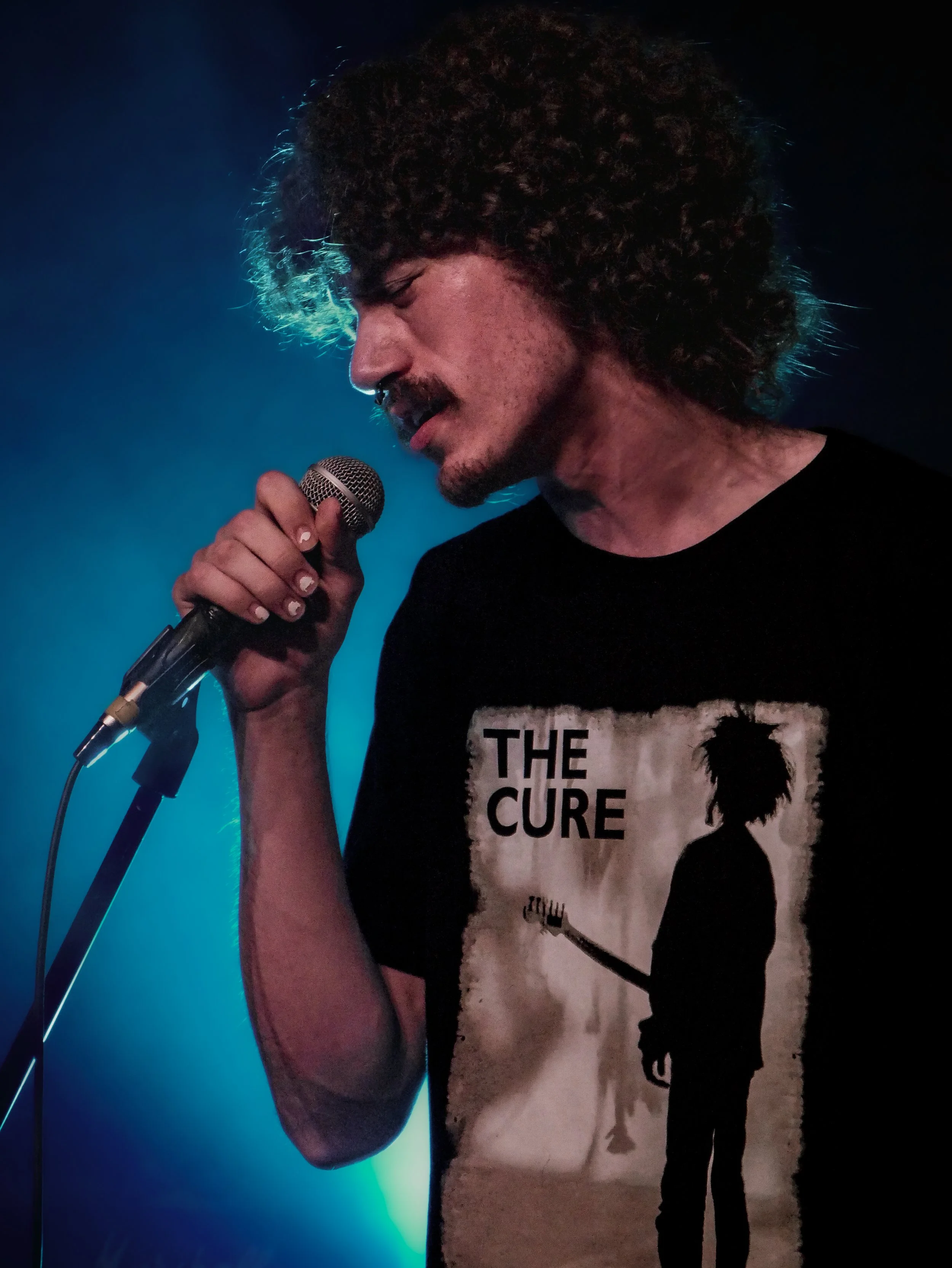 A person with curly hair and a mustache holding a microphone, wearing a black The Cure band T-shirt with a silhouette of a person with dreadlocks.