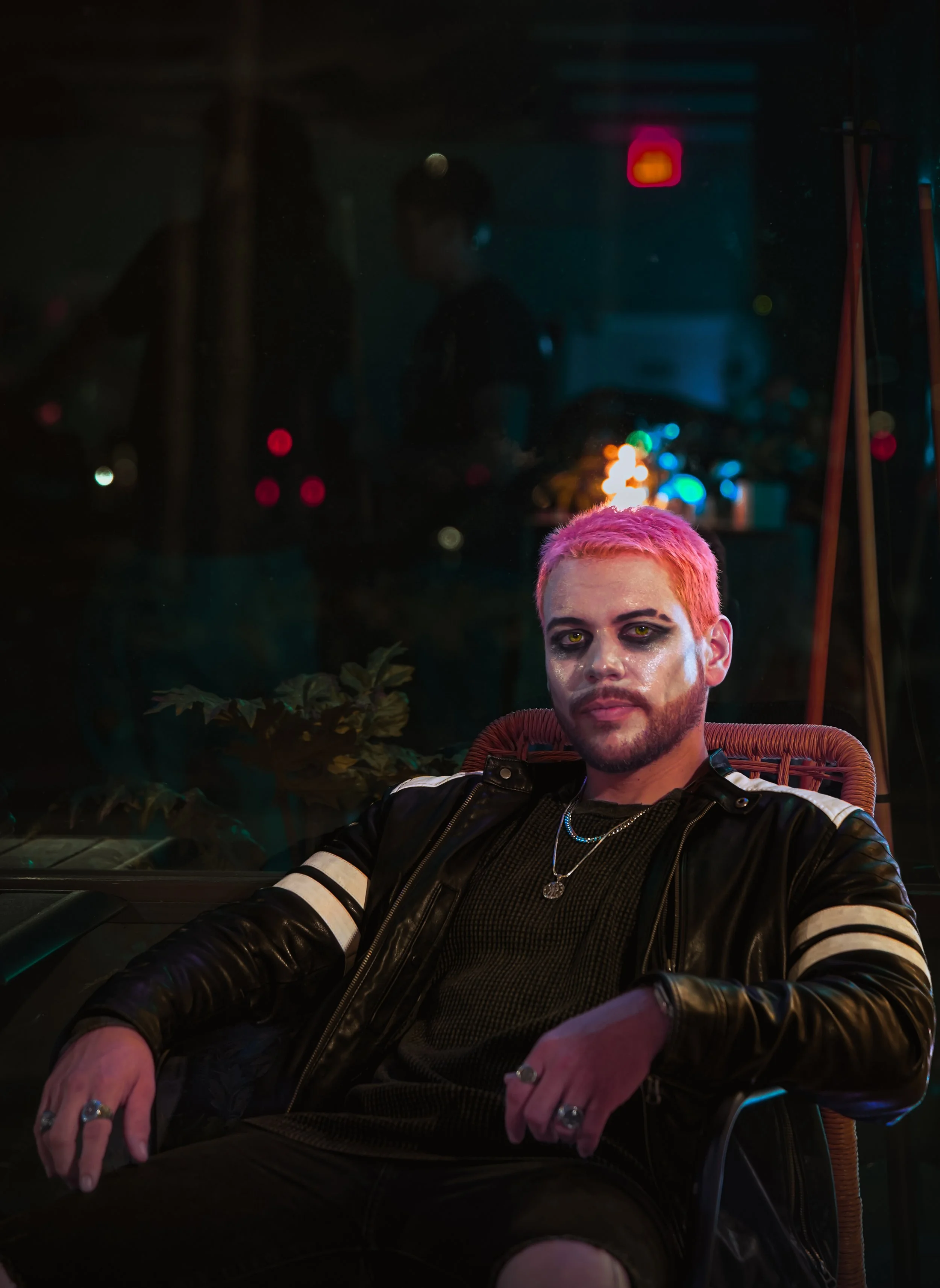 Person with pink hair and dark makeup sitting outdoors at night, wearing a black leather jacket with white stripes, jewelry, and looking into the camera, with a dark background and blurred colorful lights.