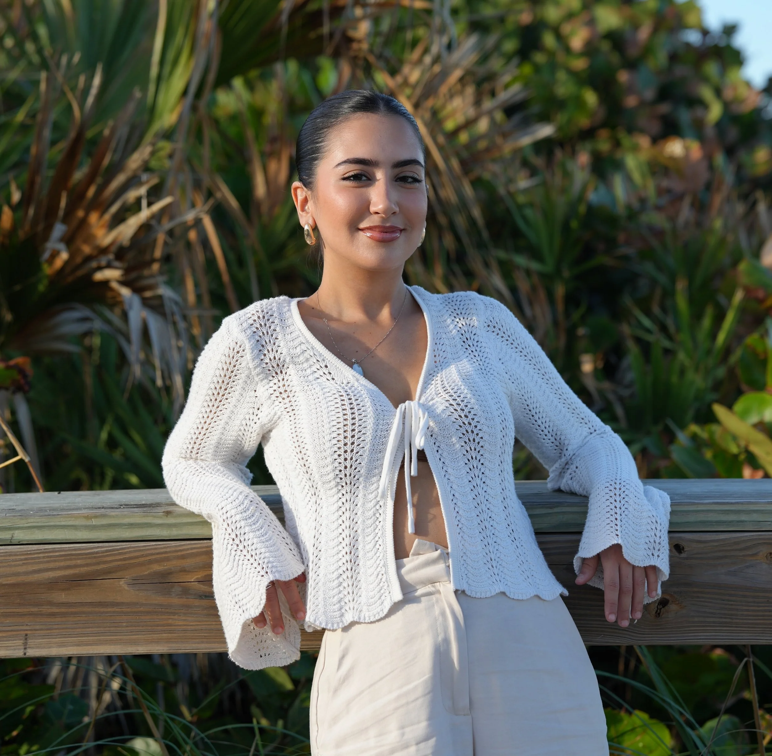 A woman with dark hair styled in a bun, wearing a white knitted cardigan, earrings, and a necklace, standing outdoors on a wooden railing with greenery and plants behind her.