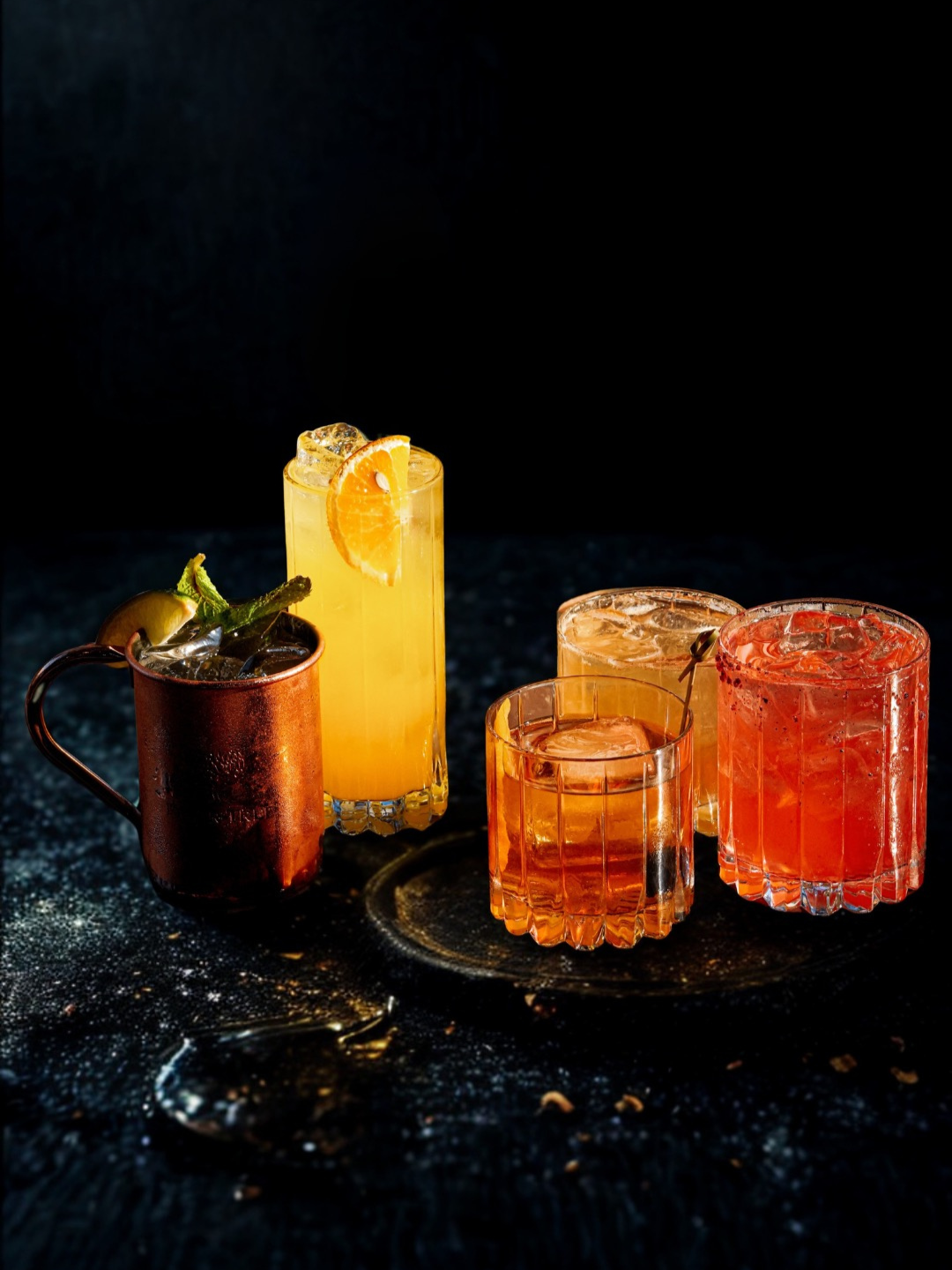 Four colorful cocktails with ice on a dark textured surface against a black background.