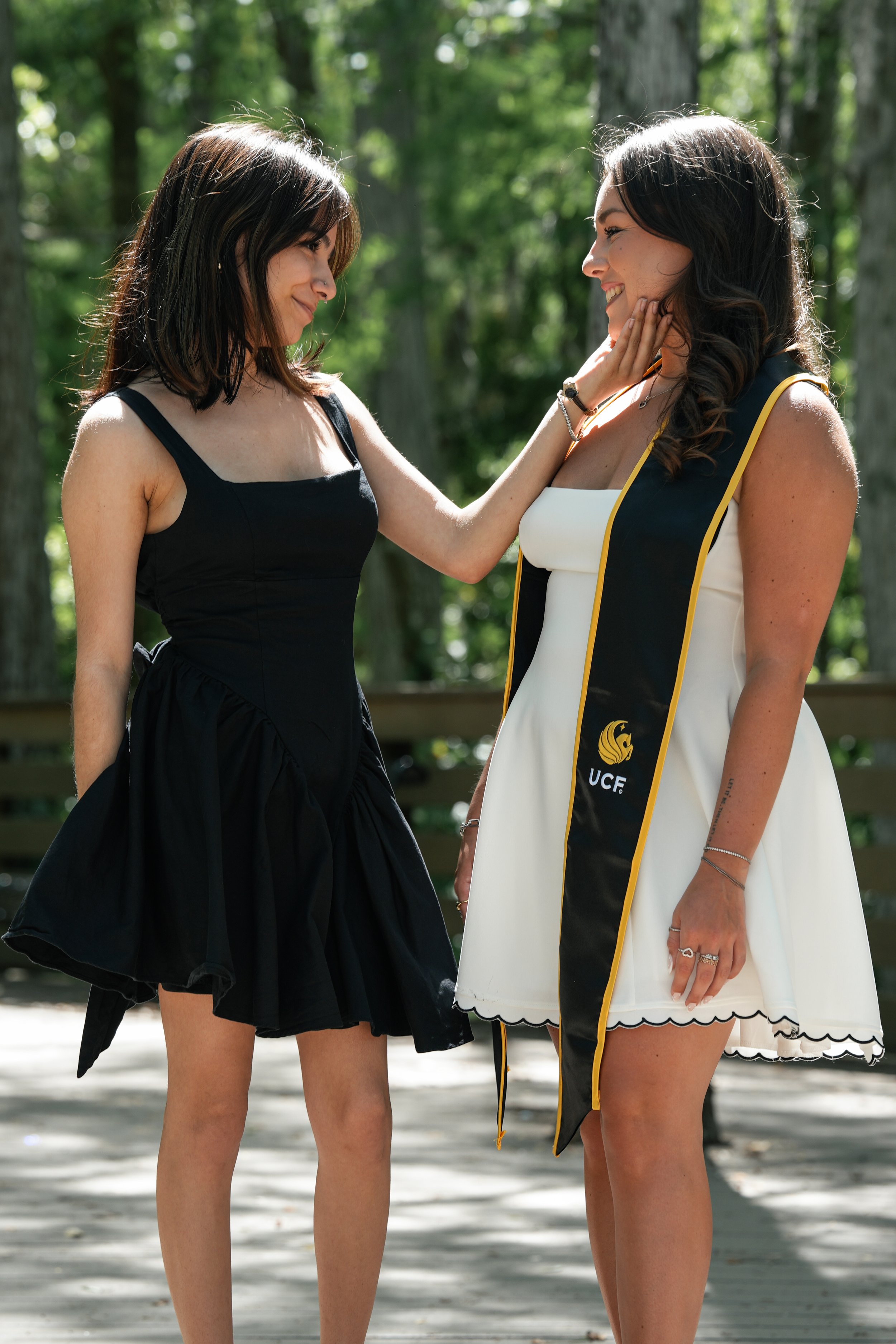 Two women, one wearing a black dress and the other wearing a graduation gown, sharing a moment outdoors with greenery in the background.