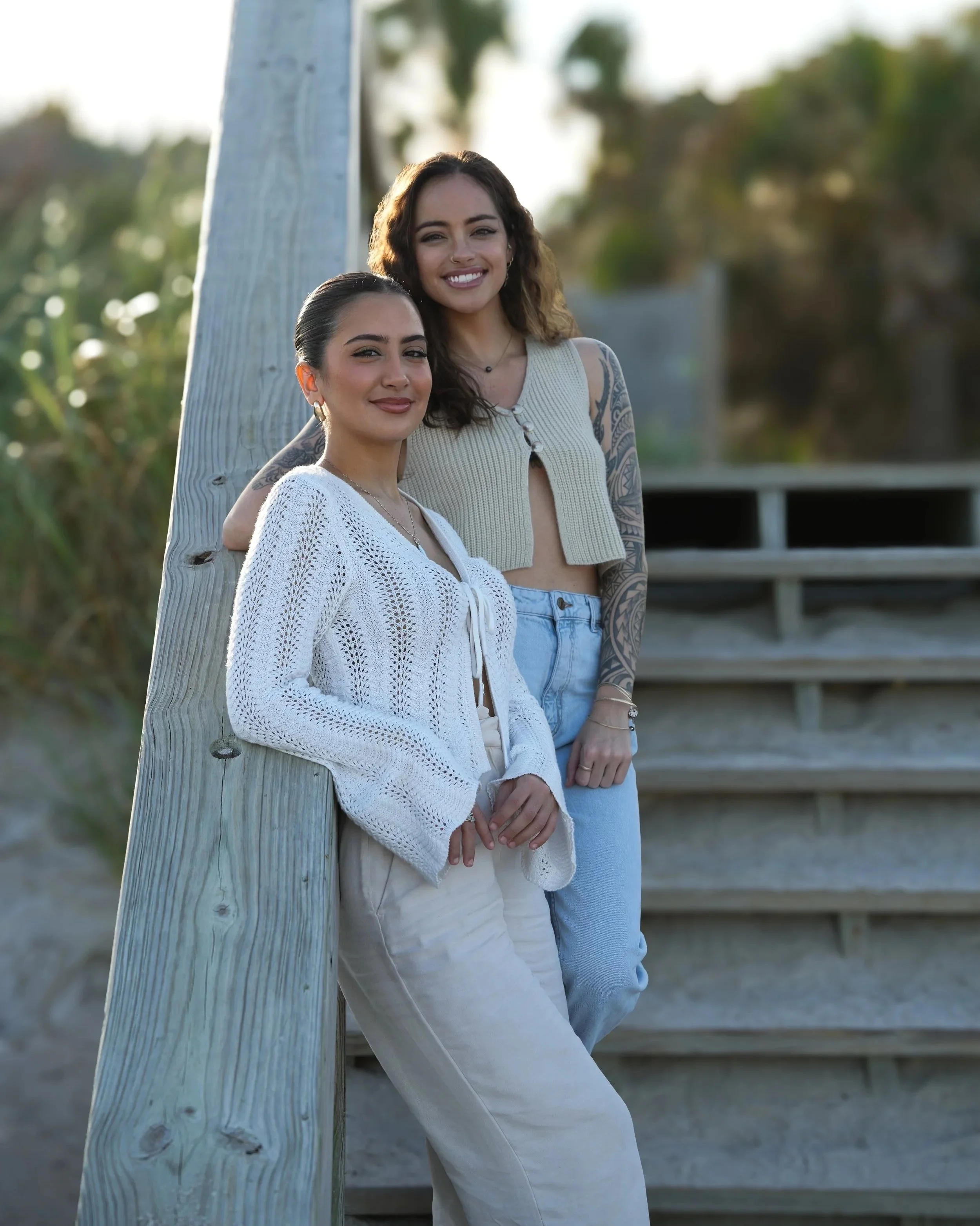 Two women standing outdoors, leaning against a wooden railing, smiling and looking at the camera, with greenery in the background.