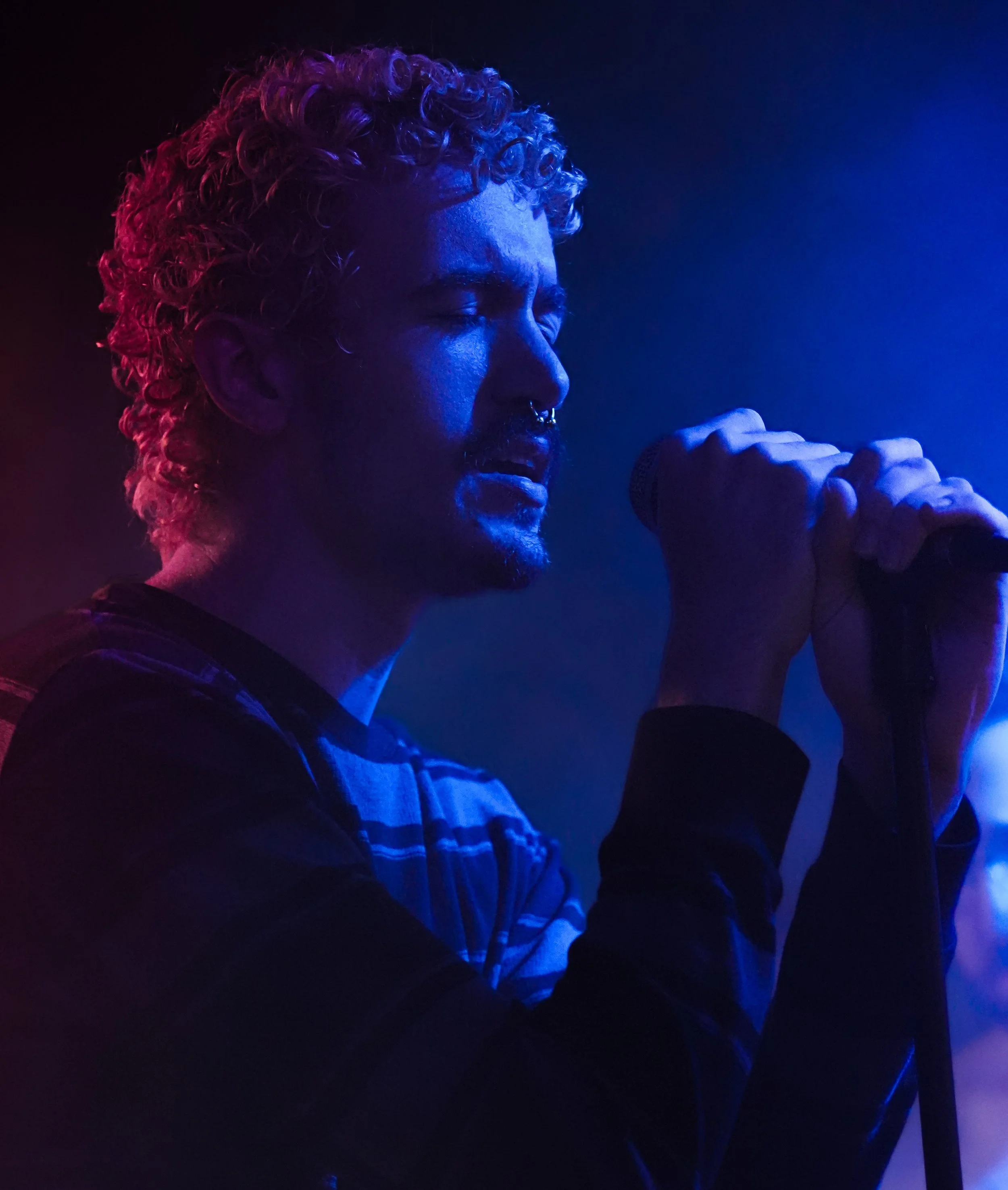 A person with curly blonde hair singing into a microphone, illuminated by blue and purple stage lighting.