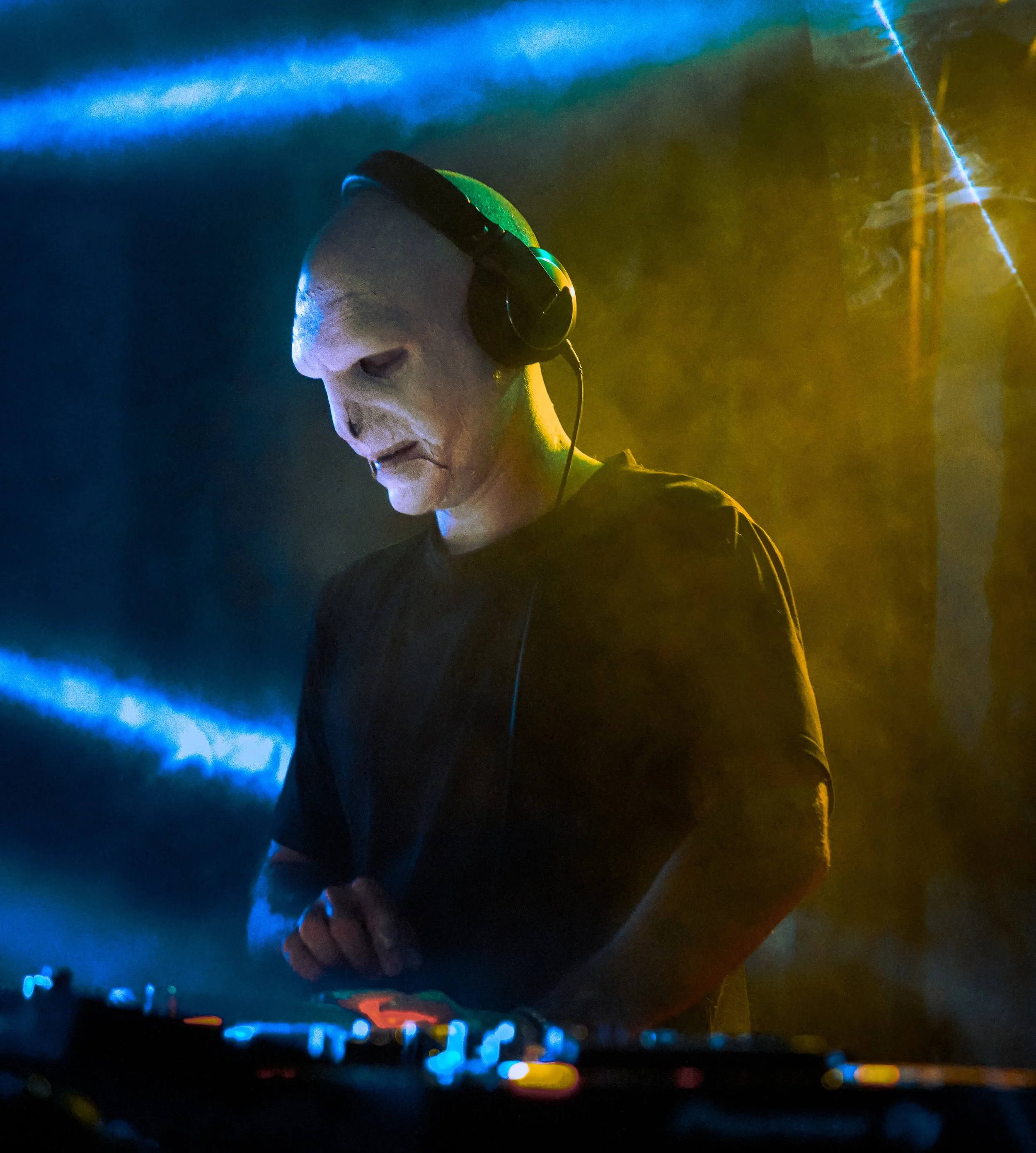A person wearing a white mask resembling a skull, with headphones on, standing behind a DJ console with colorful lights, in a dimly lit environment with yellow and blue lighting.