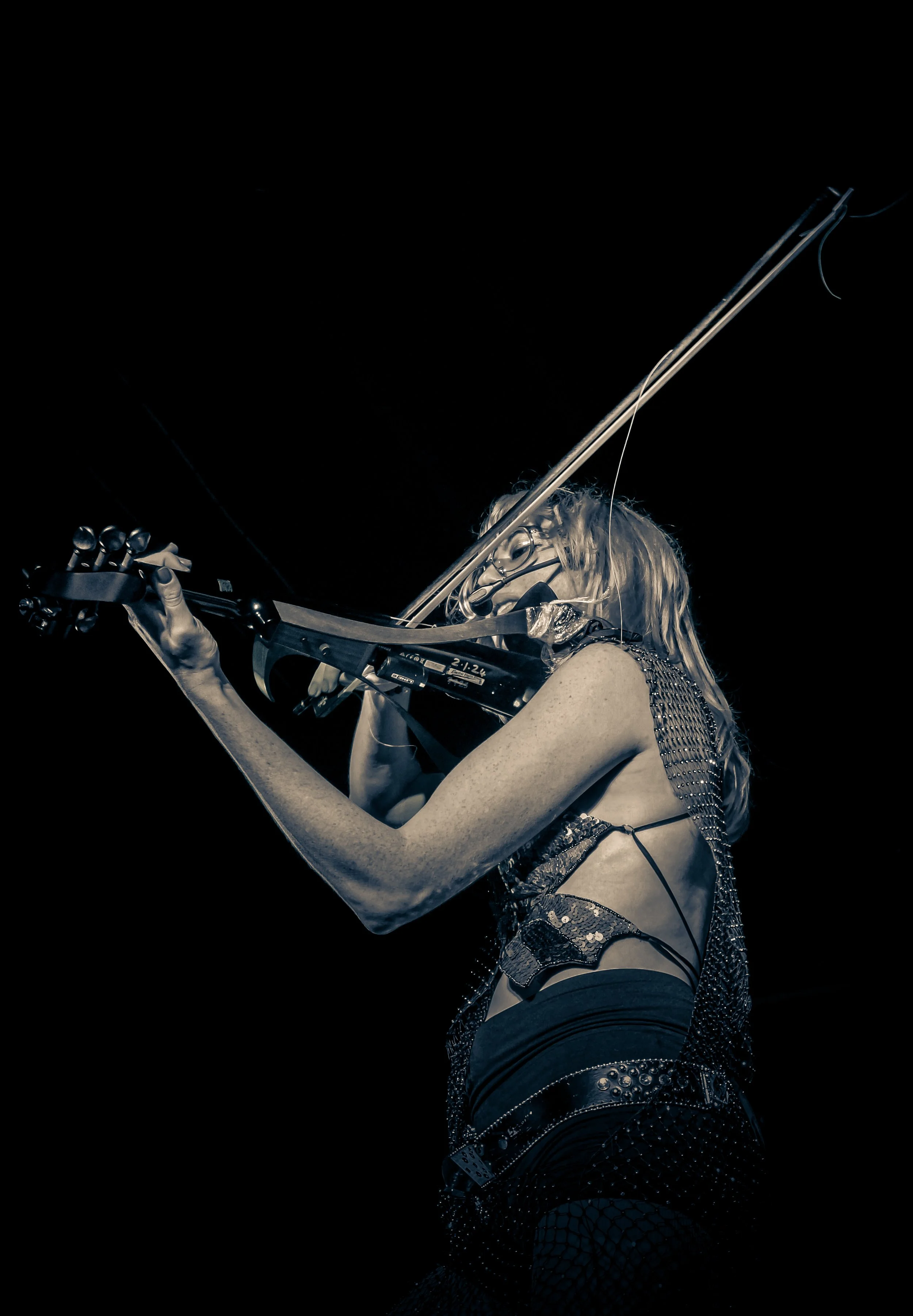 A female musician playing the violin on stage, wearing a sleeveless, metallic, and strappy outfit with fishnet pants, with a dark background.