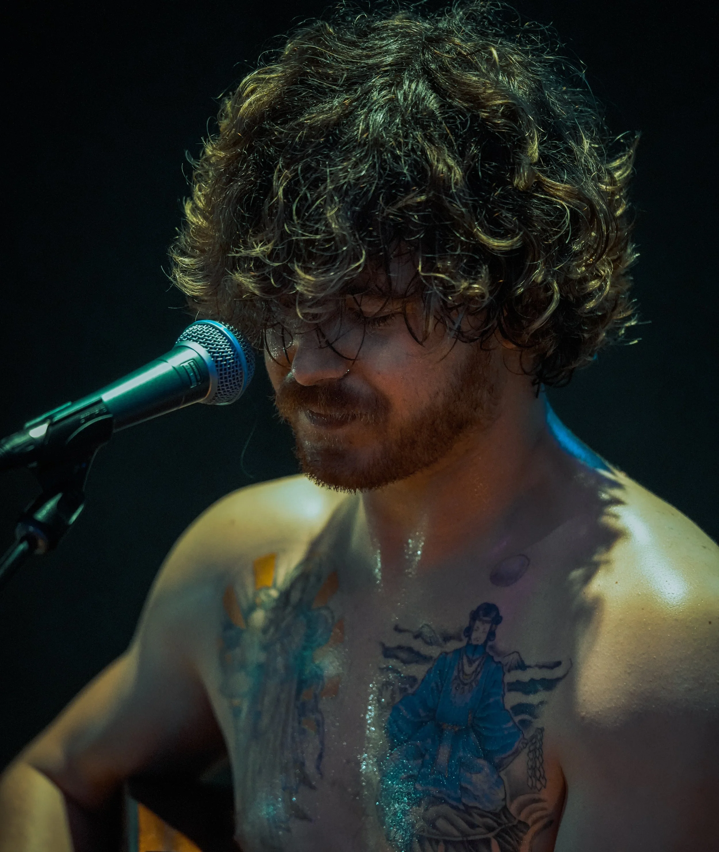 A shirtless man with curly hair and tattoos on his chest, sitting in front of a microphone, appears to be performing or recording.