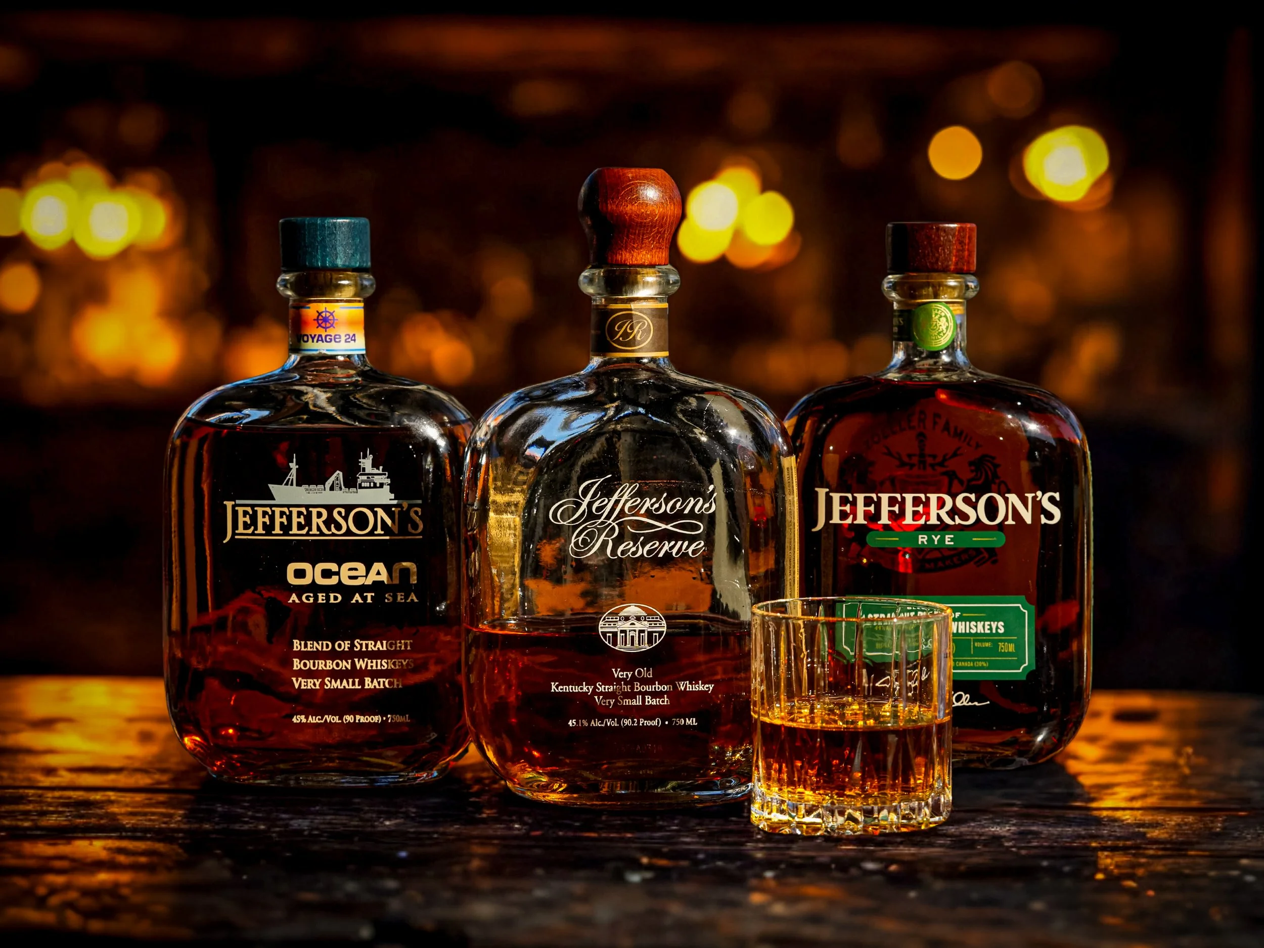 Three bottles of Jefferson's whiskey on a wooden surface with a glass filled with whiskey in front of the middle bottle, against a warm, blurred background.
