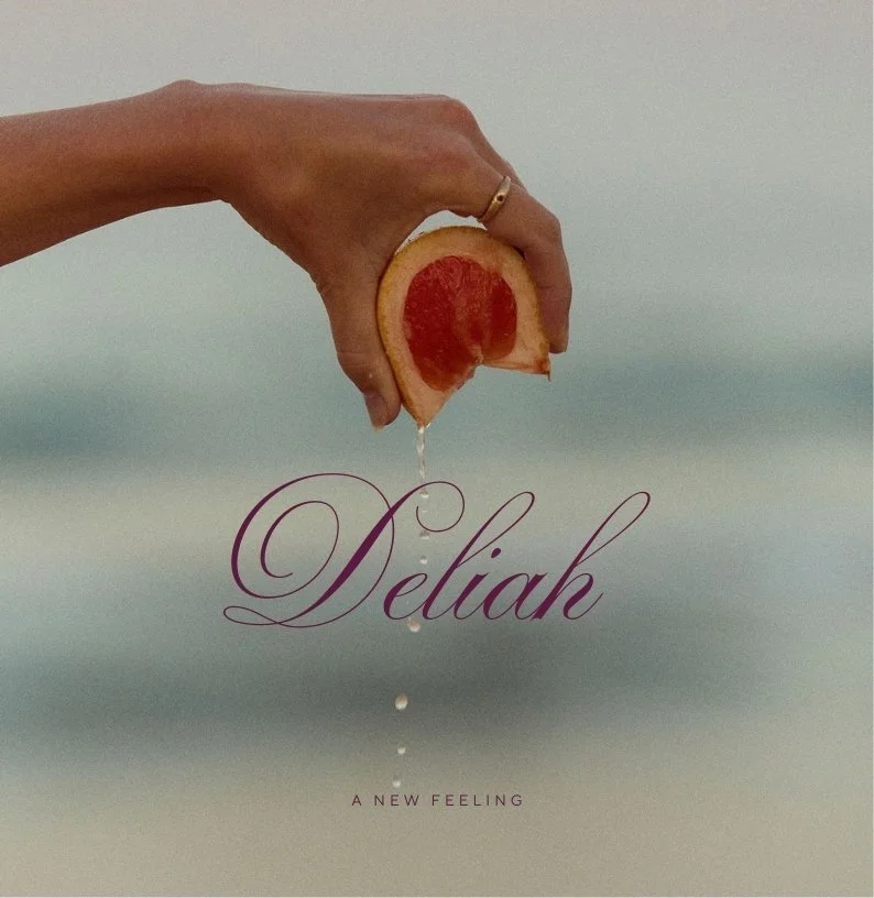 A hand holding a slice of grapefruit with water droplets falling from it, with the word 'Deliah' written in elegant script and the phrase 'A New Feeling' below.