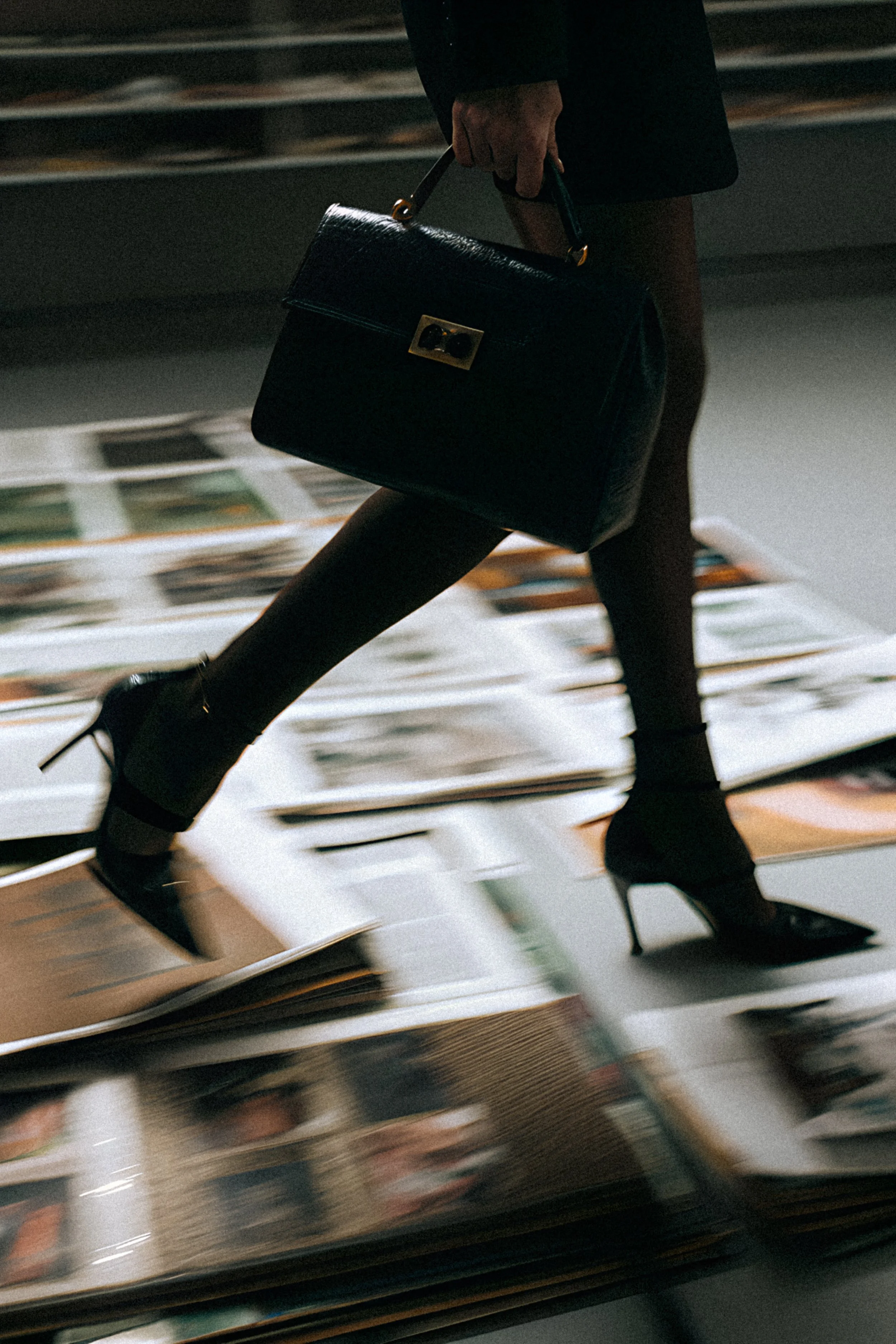 A person walking with a black handbag, black high-heeled shoes, and black tights in an environment with blurred magazines or papers on the floor.