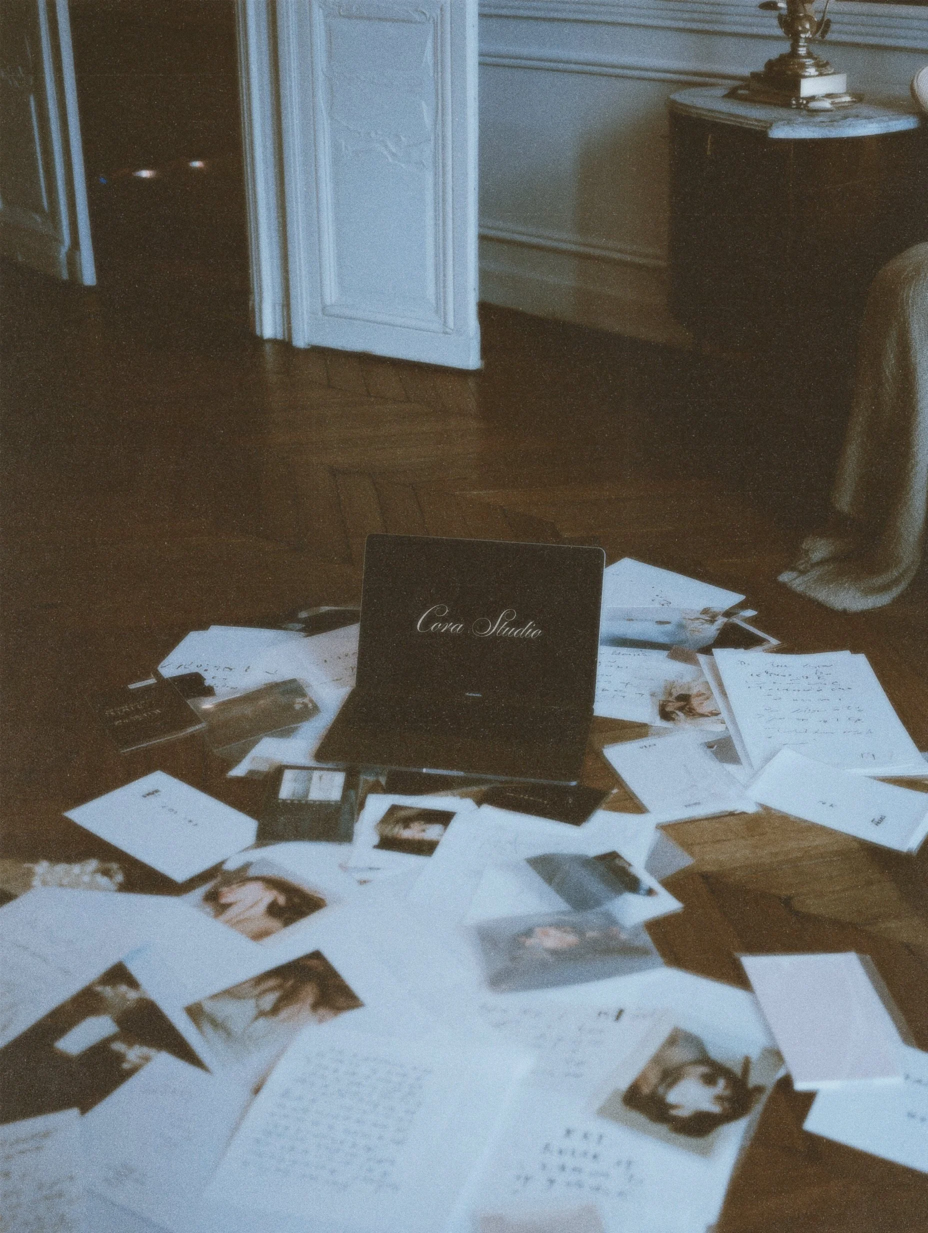 Disorganized papers, photographs, and a laptop on a wooden floor in a room with white walls and a black side table.