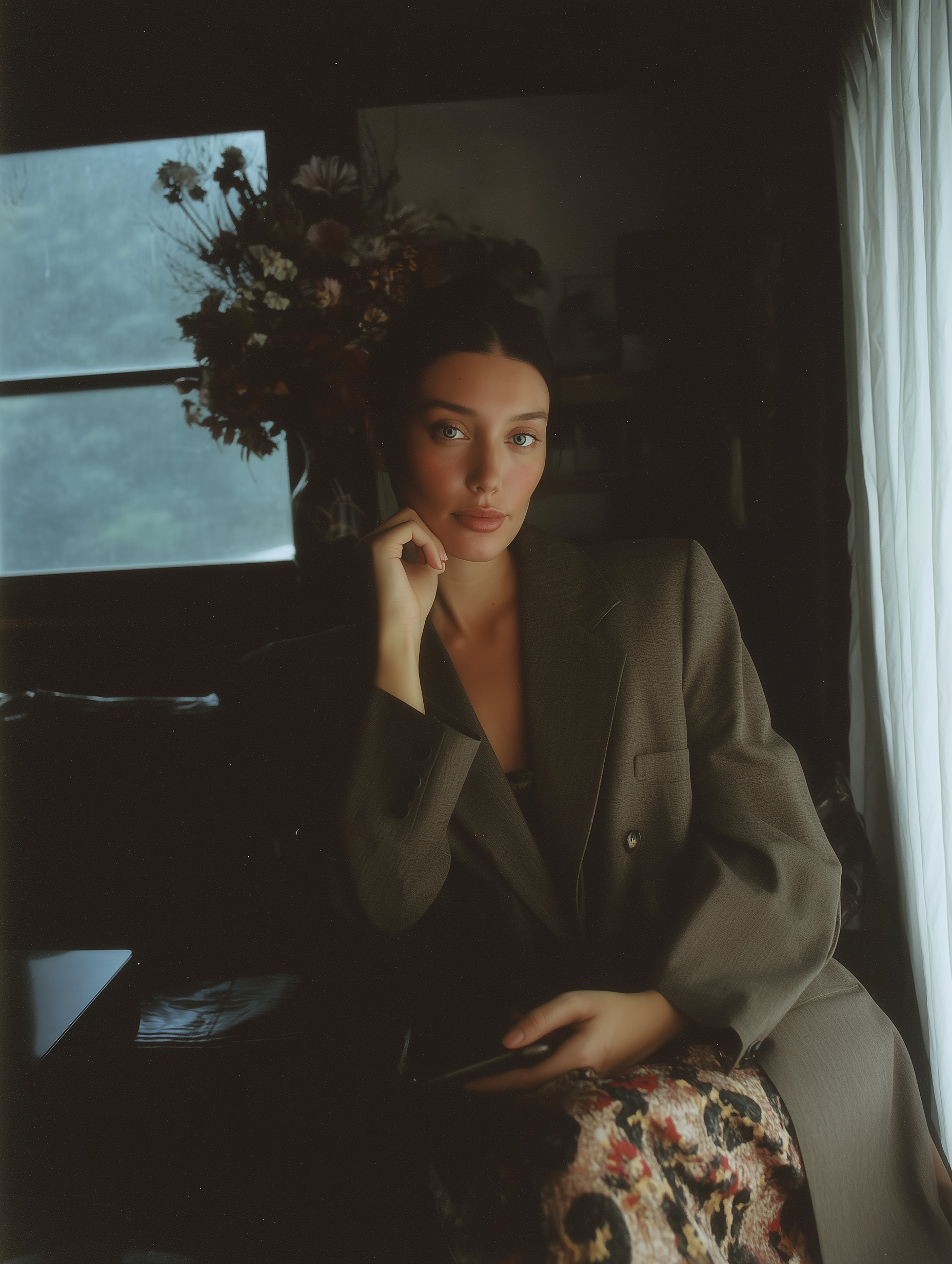 A woman with dark hair and blue eyes is sitting indoors near a window, resting her chin on her hand. She is wearing a brown blazer and has a floral garment partially visible. Behind her, there is a large floral arrangement on her head.