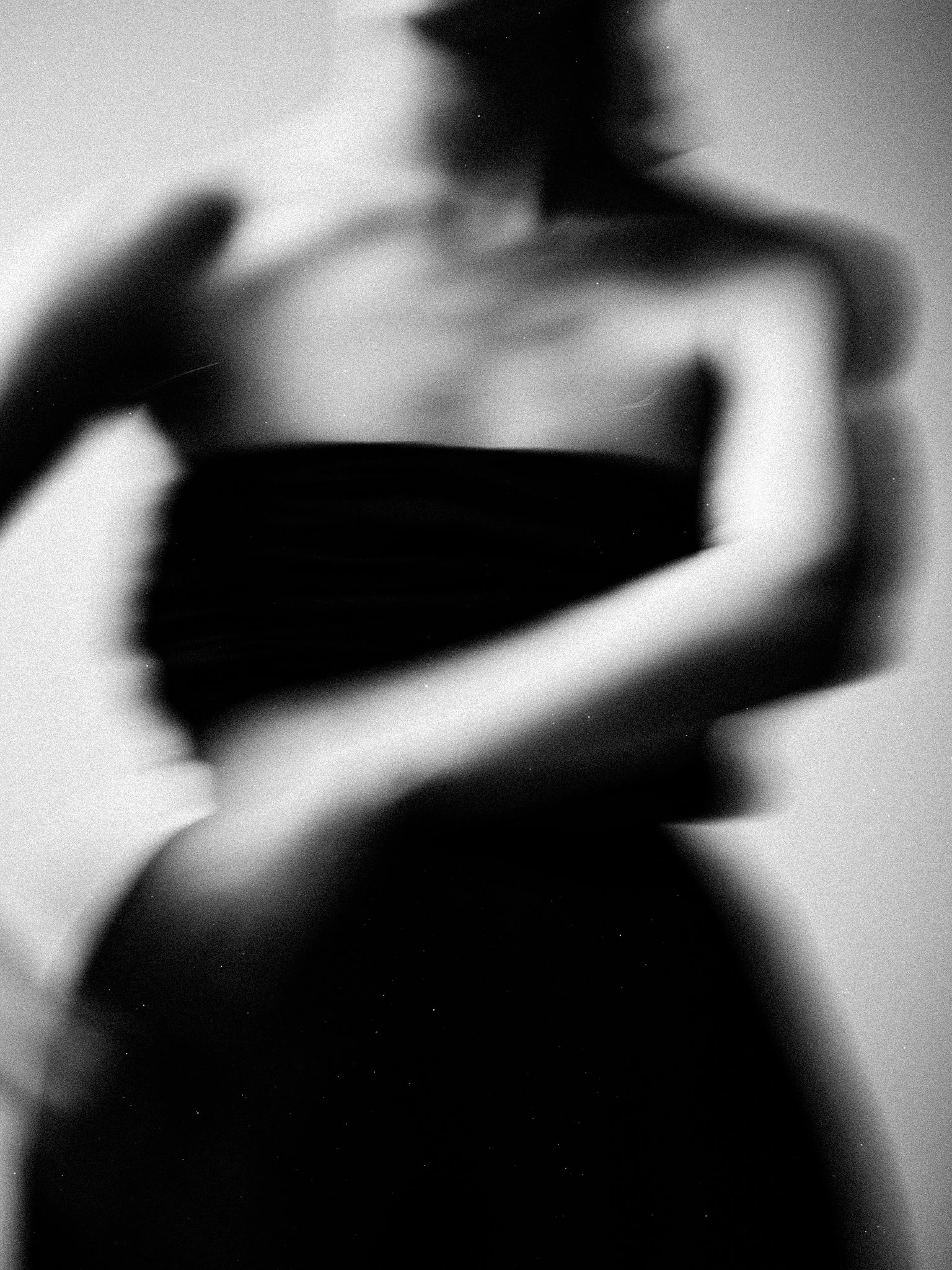A black and white, blurry photo of a person with shoulder-length hair, wearing a strapless top, with one hand resting on their shoulder and the other across their abdomen, in a minimalistic pose.