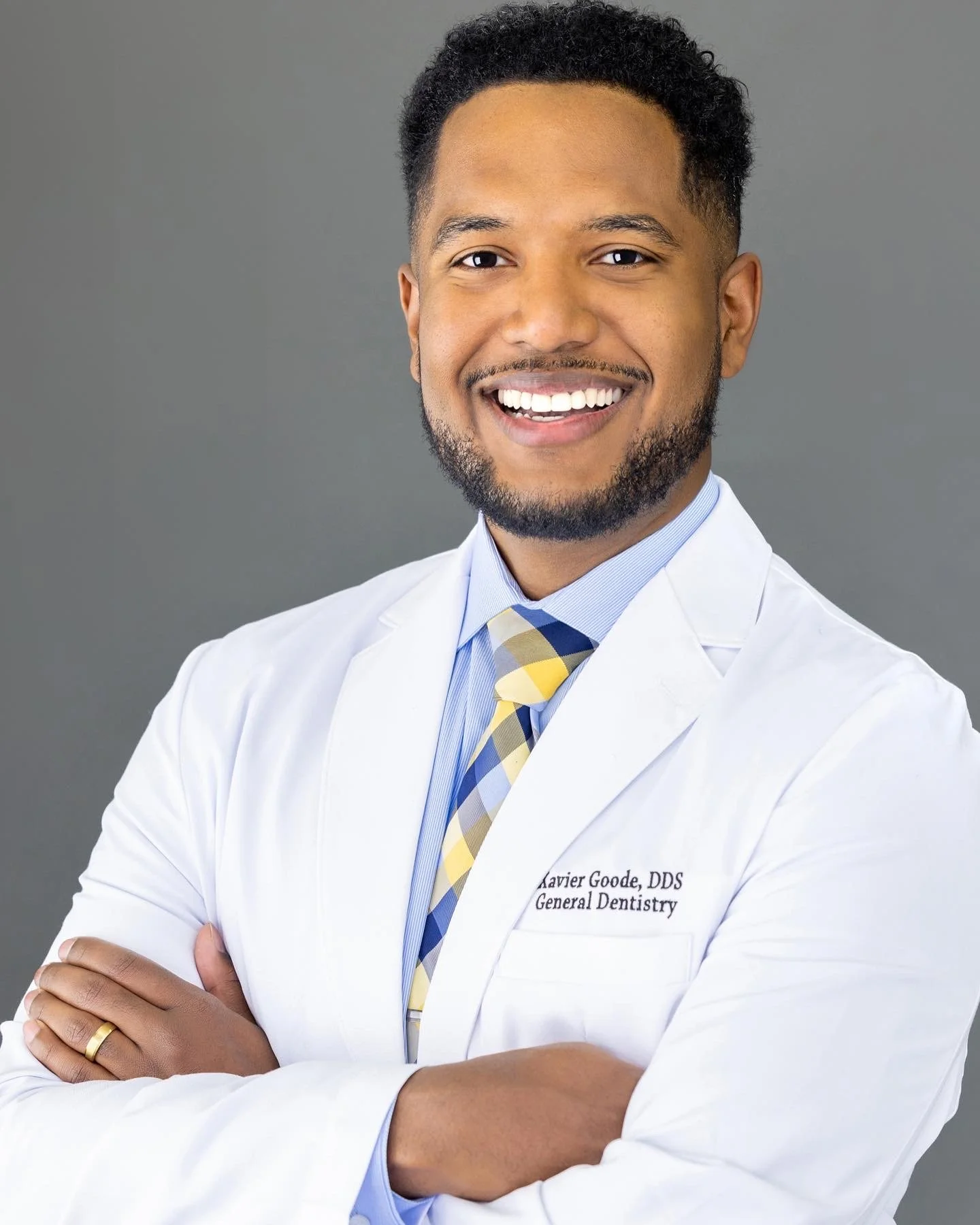 A smiling man in a white doctors coat with crossed arms, wearing a blue shirt and yellow plaid tie, standing against a gray background.