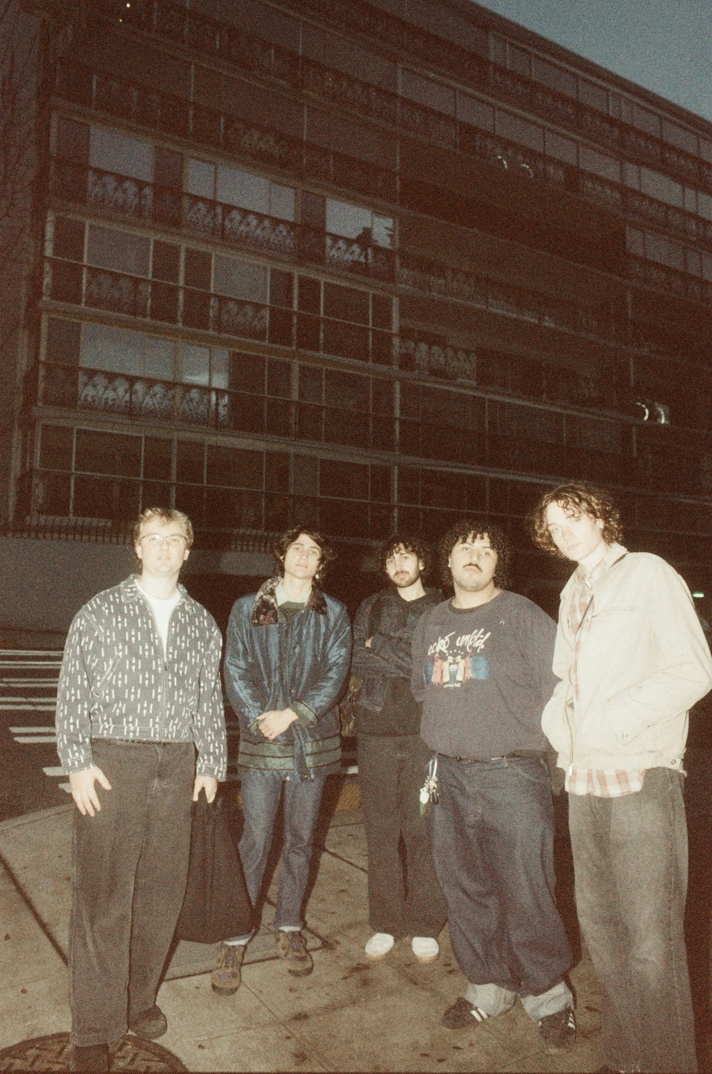 Five young men standing outside at night in front of a multi-story building with many windows and balconies. Musical artist band seattle indie rock rnb slacker alt-country independent music band pictures. Jackie Tulsa is a music band.