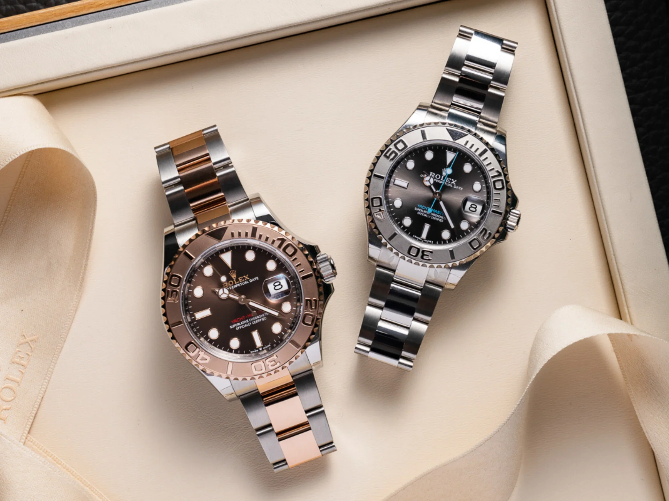 Two Rolex watches in a beige box, one with a black dial and rose gold bezel, the other with a black dial and silver bezel, both with metal link bracelets.