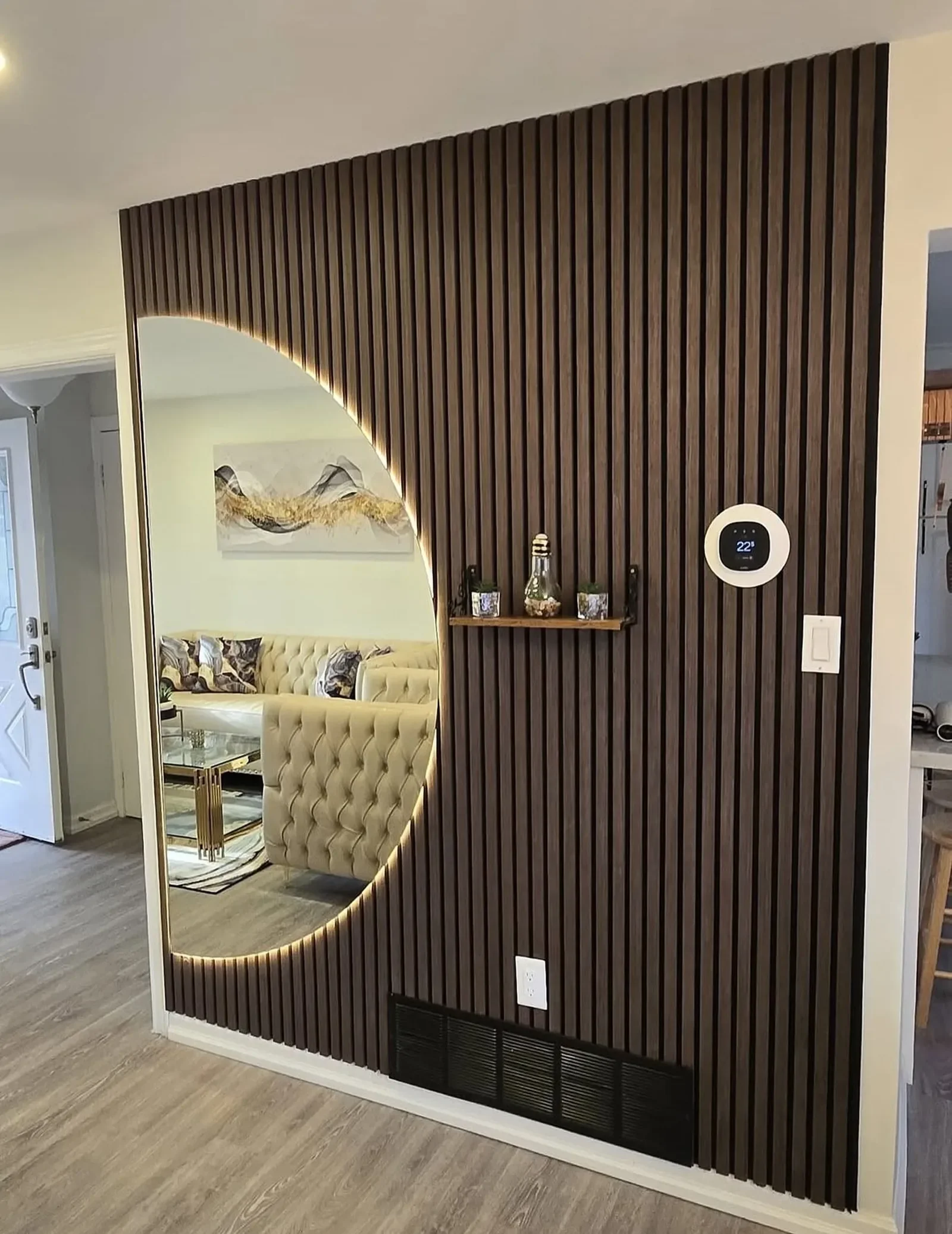 Modern living room viewed through a decorative wooden slat wall with LED lighting, featuring a mirror, small shelf with decor, and thermostat display.