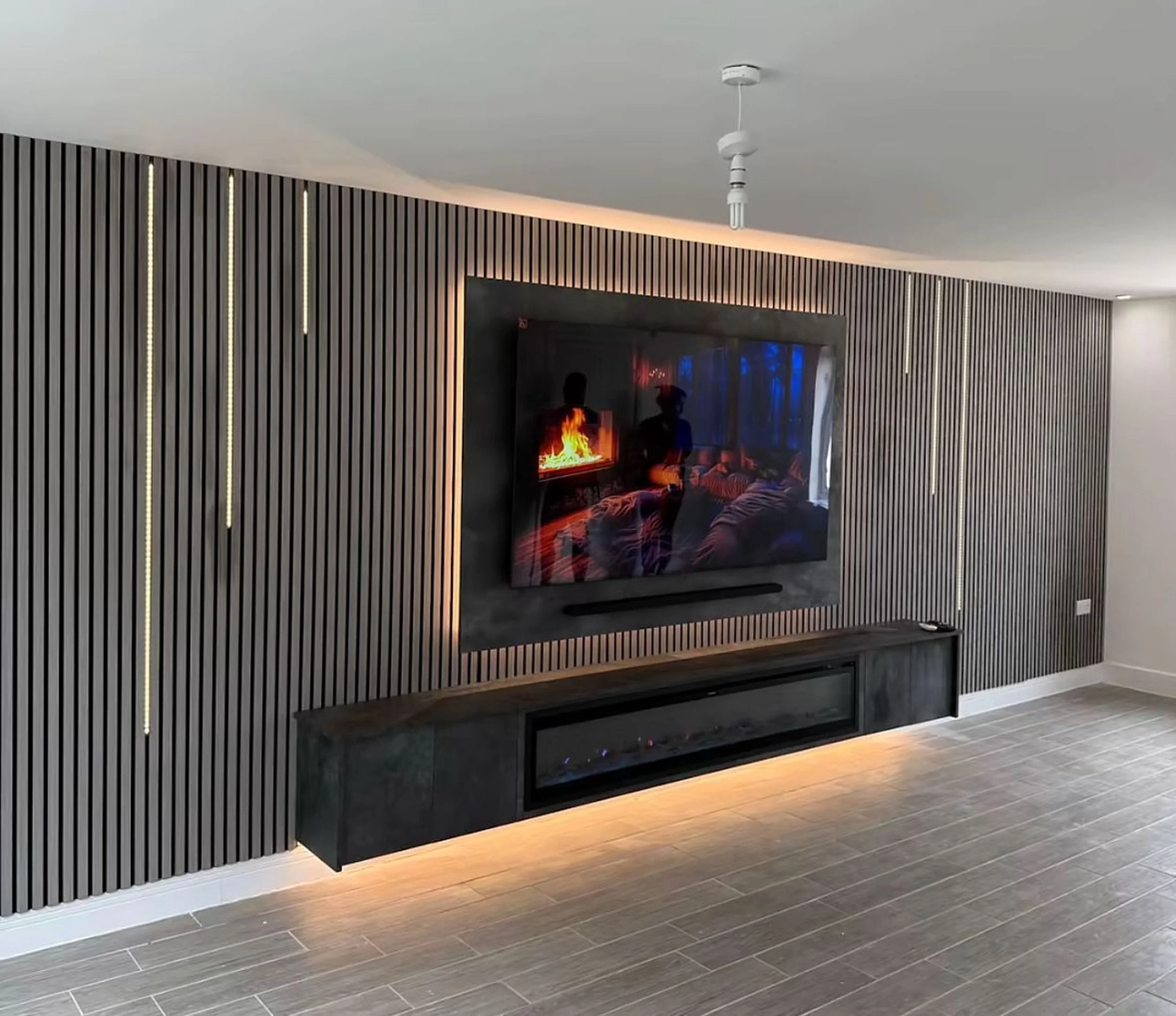 Modern living room with a wall-mounted flat-screen TV, a black wooden cabinet underneath, and a fireplace reflected on the TV screen, with vertical black wooden slats and LED lighting accents on the wall, and wooden flooring.
