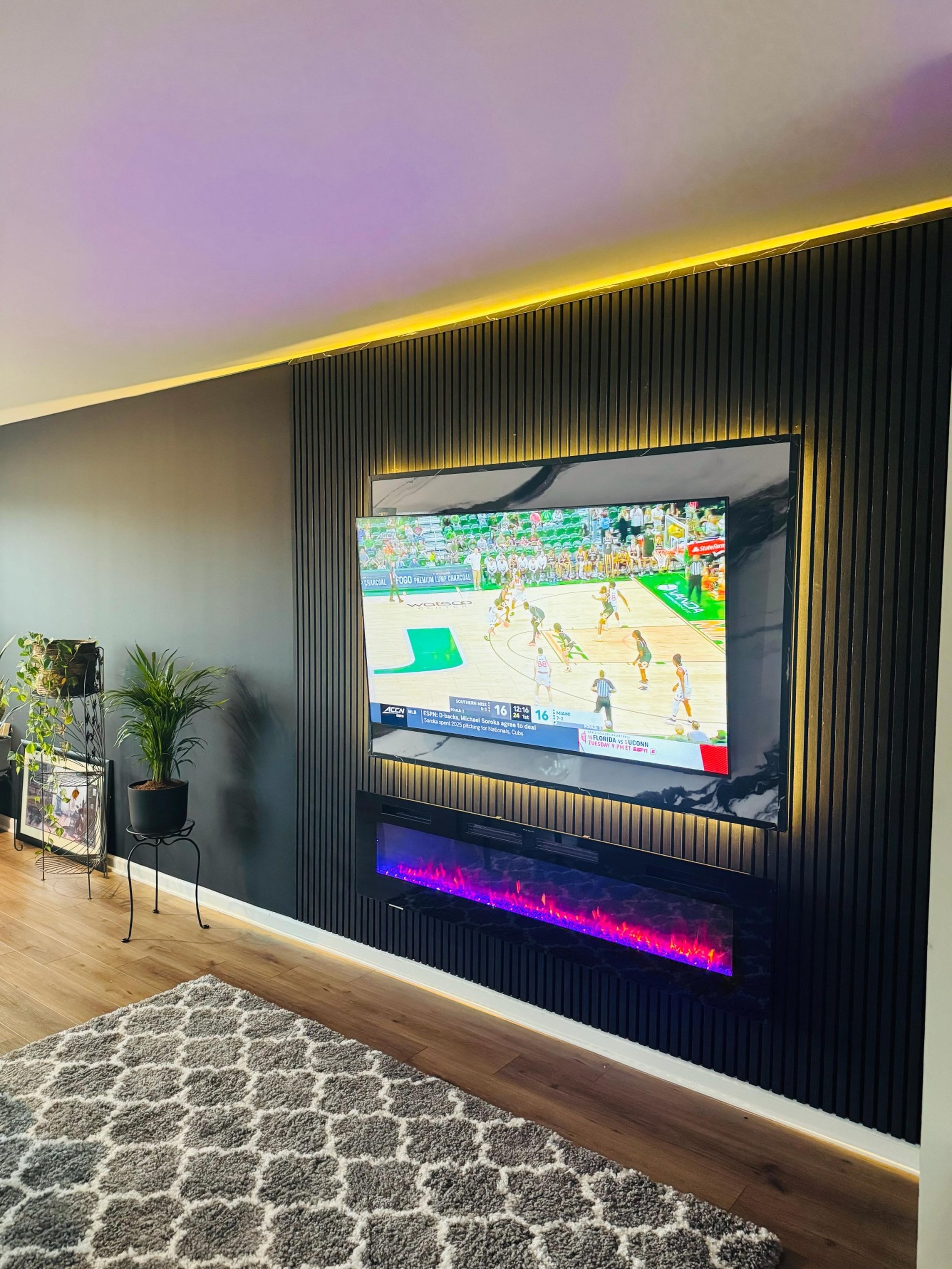 Living room with a wall-mounted flat-screen TV showing a basketball game, black textured wall paneling, an electric fireplace with purple flames, potted plants, a patterned gray rug, and hardwood flooring.